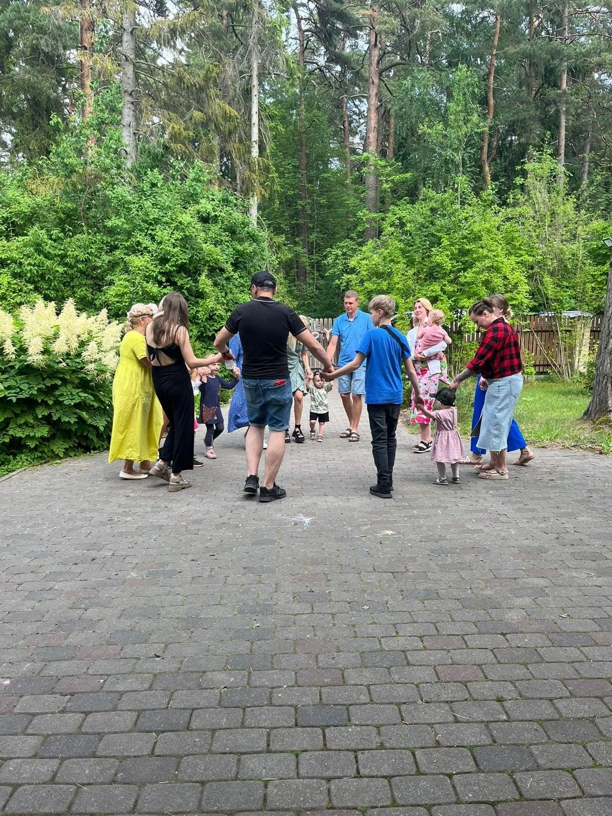 Group of children and adults holding hands in a circle outdoors on a paved path surrounded by green trees and bushes.