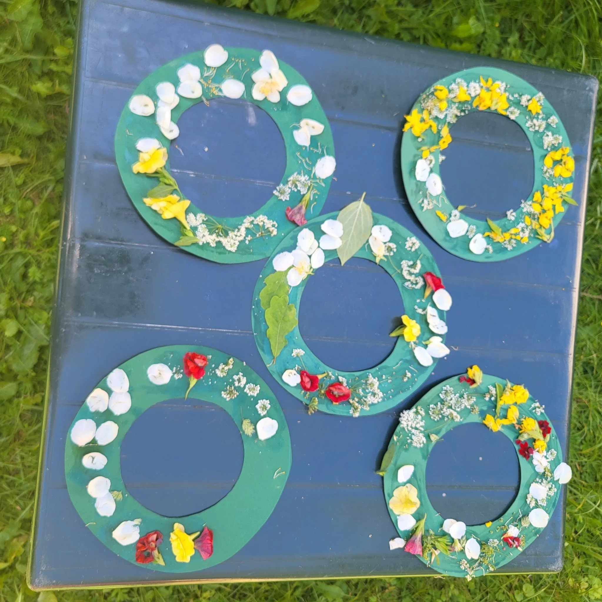 Five green circular paper wreaths decorated with various flowers and leaves, placed on a blueish wooden surface outdoors.