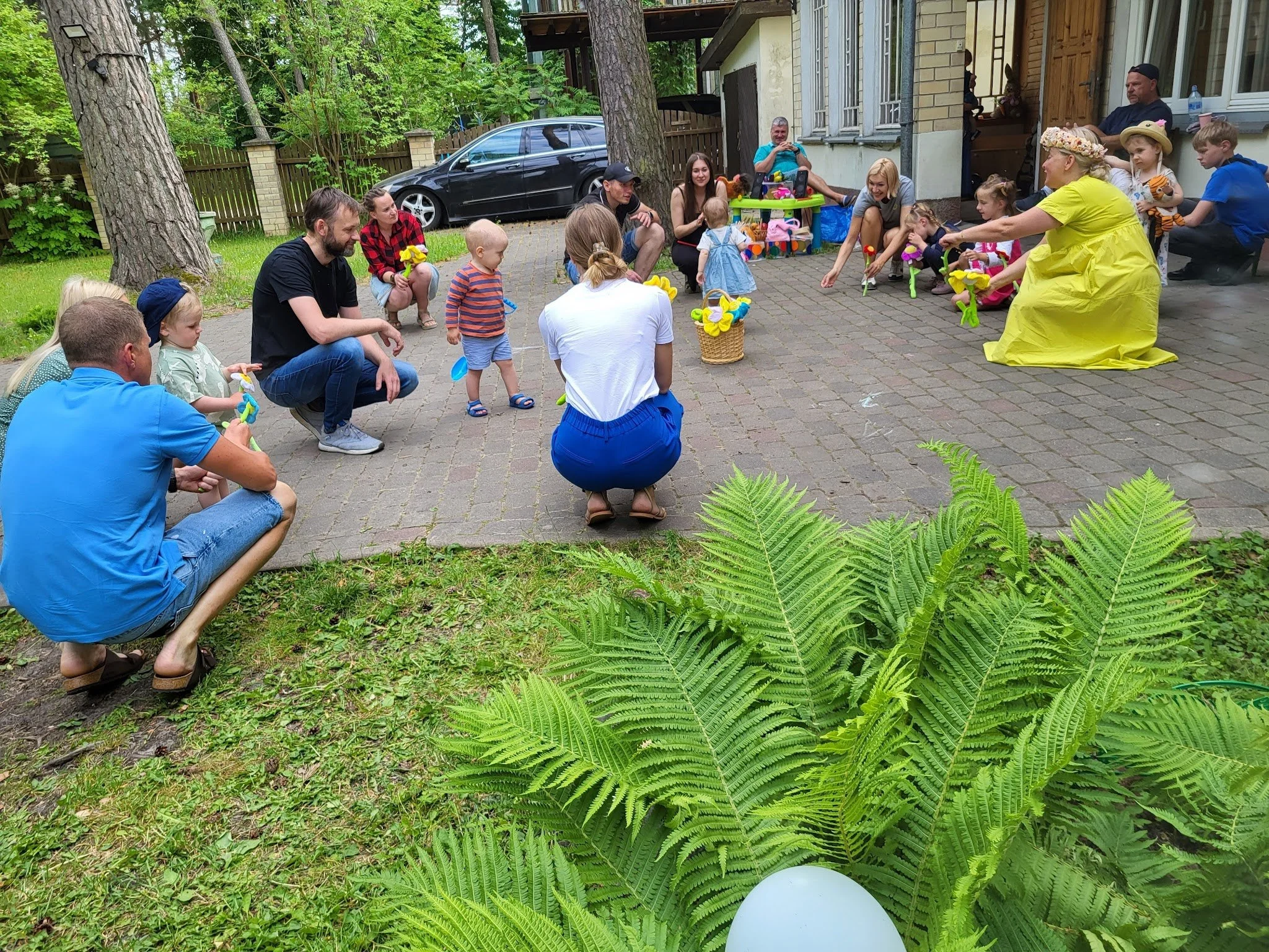 Children and adults participating in a children's Easter egg hunt in a backyard, with a woman in a yellow dress and flower hat handing out eggs, and a group of children gathering to find eggs, surrounded by trees and a house.