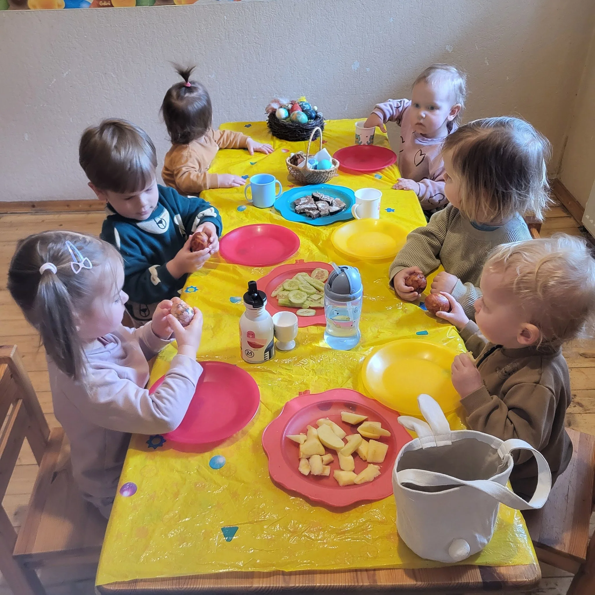Children sitting around a yellow table with pink and yellow plates, eating snacks, with a basket of decorated eggs and a plate of cookies on the table.
