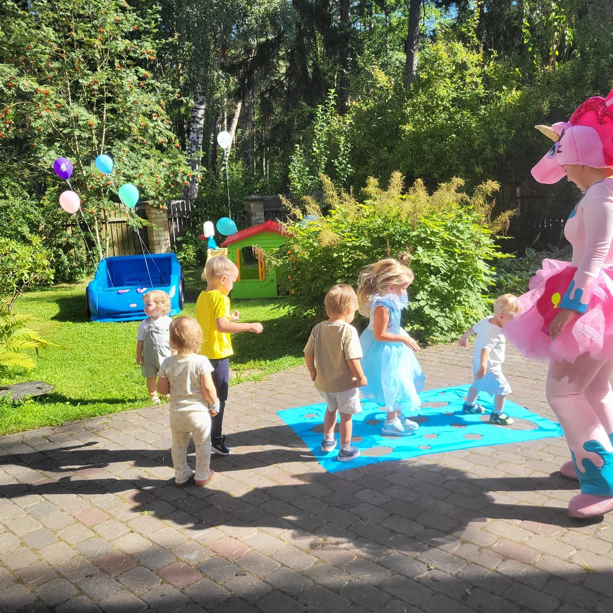 Children playing on a blue Twister mat outdoors, with a person dressed as a pink unicorn mascot watching, surrounded by greenery and balloons.