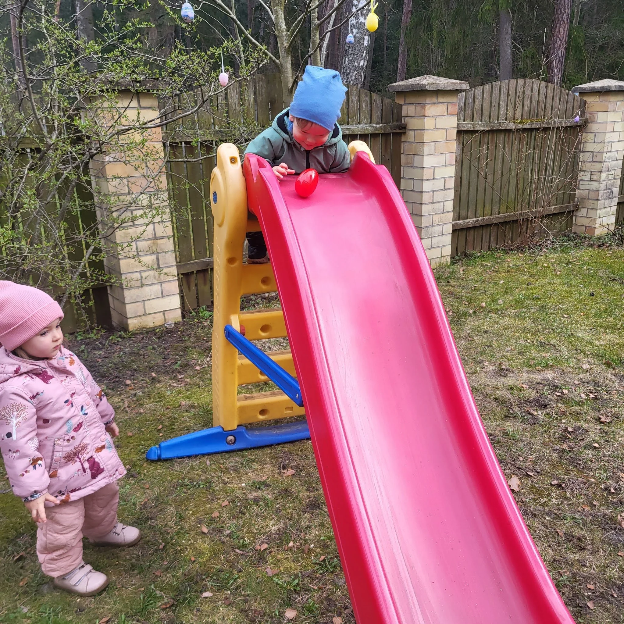 Two children outdoors near a small slide, with one child at the top holding a red Easter egg, while a girl wearing a pink jacket and hat watches.