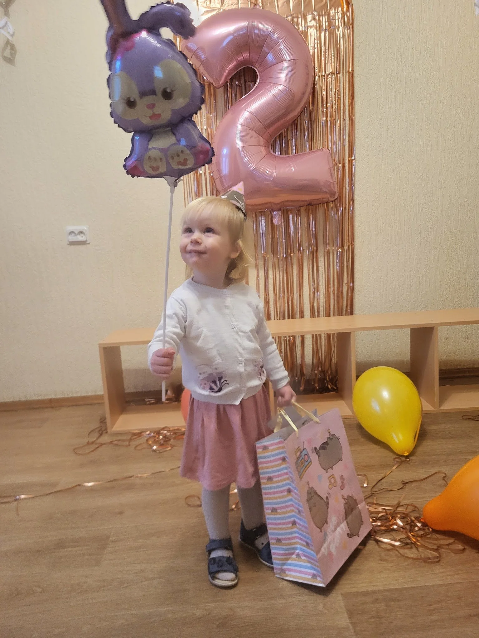 A young girl celebrating her second birthday, holding a purple balloon with a bunny illustration, a pink gift bag, and standing in front of large pink number '2' balloons, with yellow and orange balloons on the floor and a shiny pink curtain backdrop