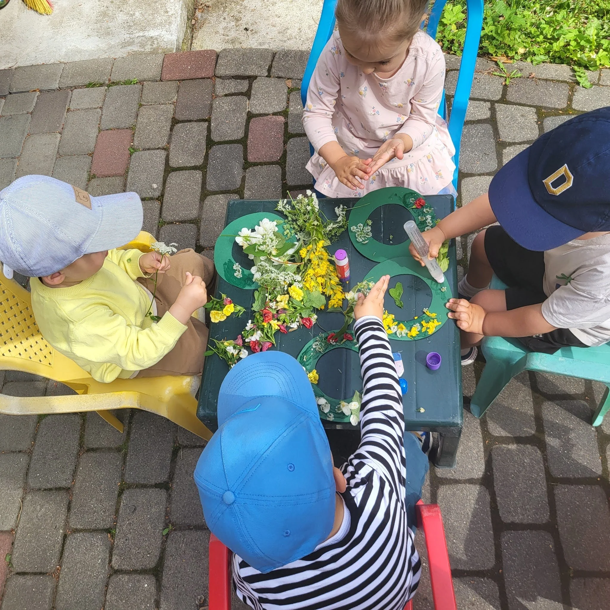 Four children sitting around a small table outdoors, working on a craft project with colorful flowers and green paper cutouts.