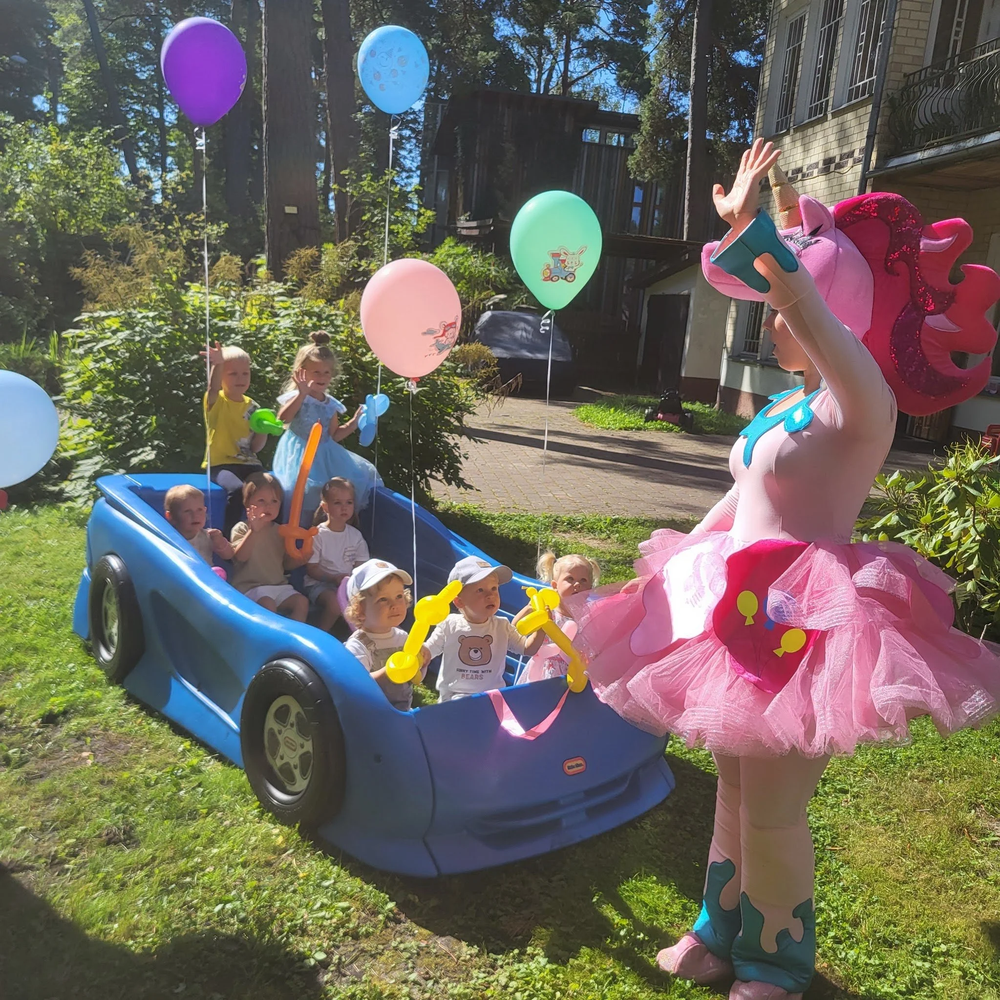 Children sitting in a blue toy car holding balloons with a person dressed as a colorful unicorn or fairy princess in a pink tutu and unicorn headpiece at an outdoor birthday celebration.