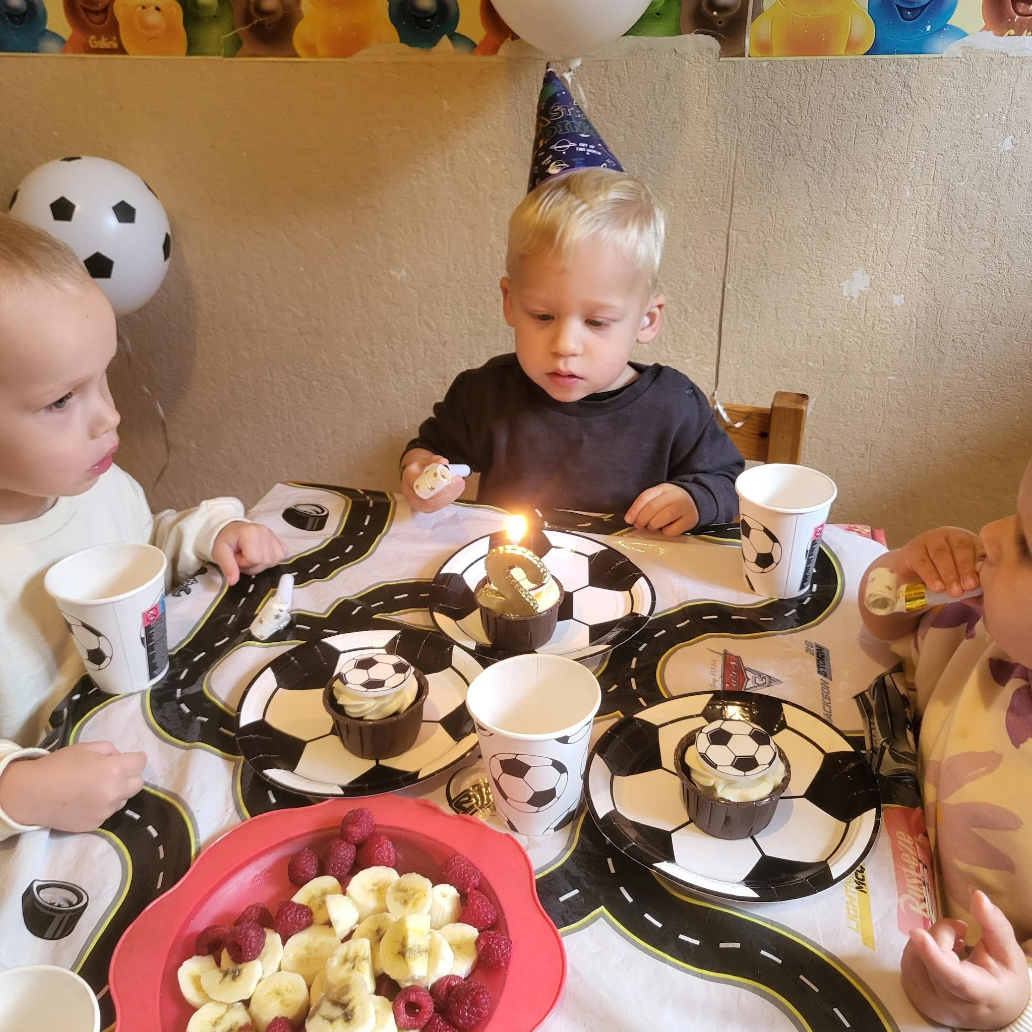 Children celebrating a birthday with a soccer-themed party, cupcakes, fruit, and party hats.