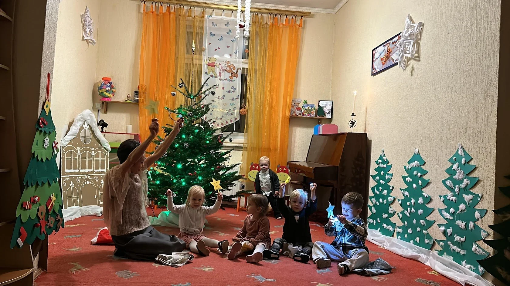 Children gathered around a decorated Christmas tree in a festive room, with holiday decorations including paper Christmas trees and a gingerbread house. An adult is sitting on the floor leading a holiday activity.