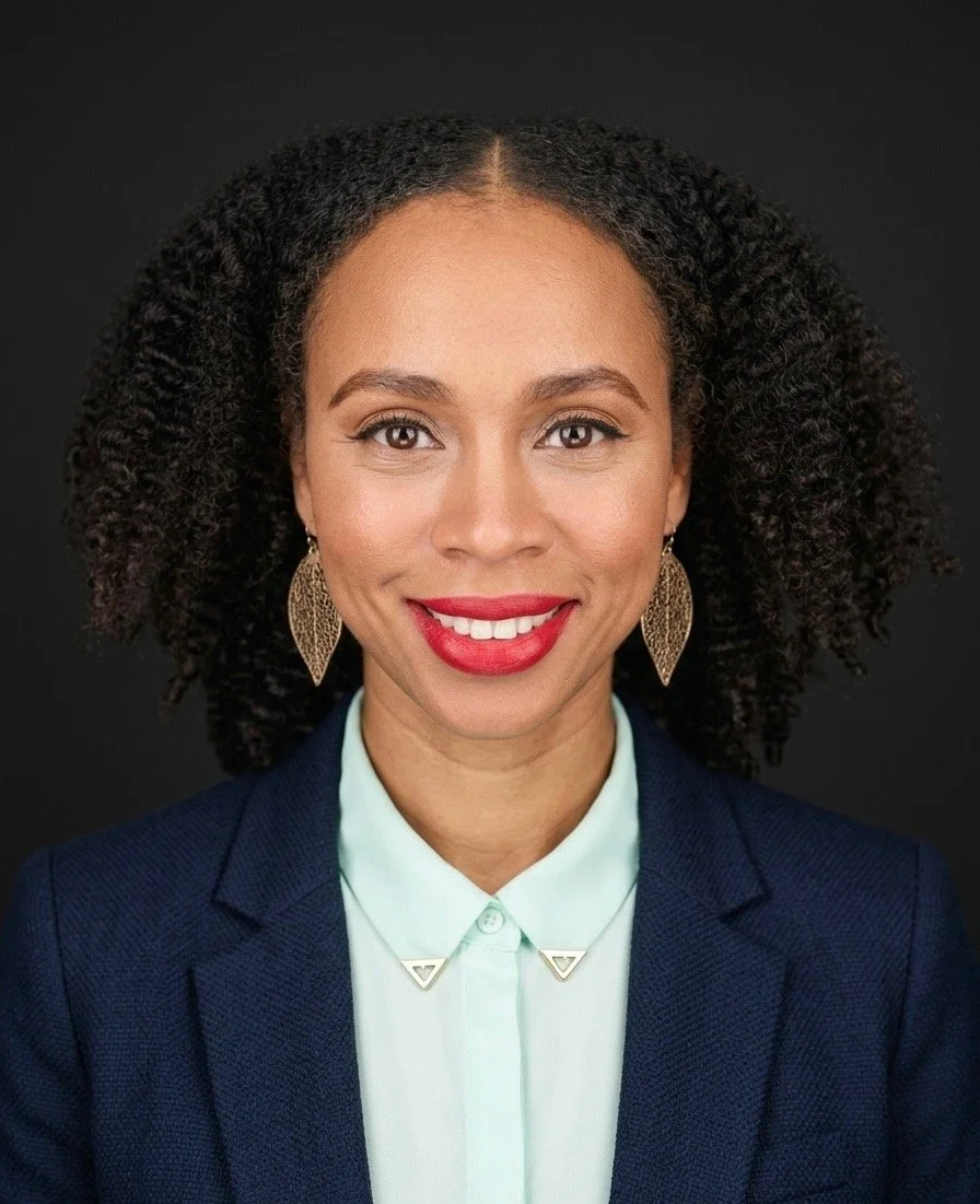 A woman (India Tizol)  with brown hair and glasses, smiling brightly, wearing a black leather jacket, standing against a textured, orange brick wall.