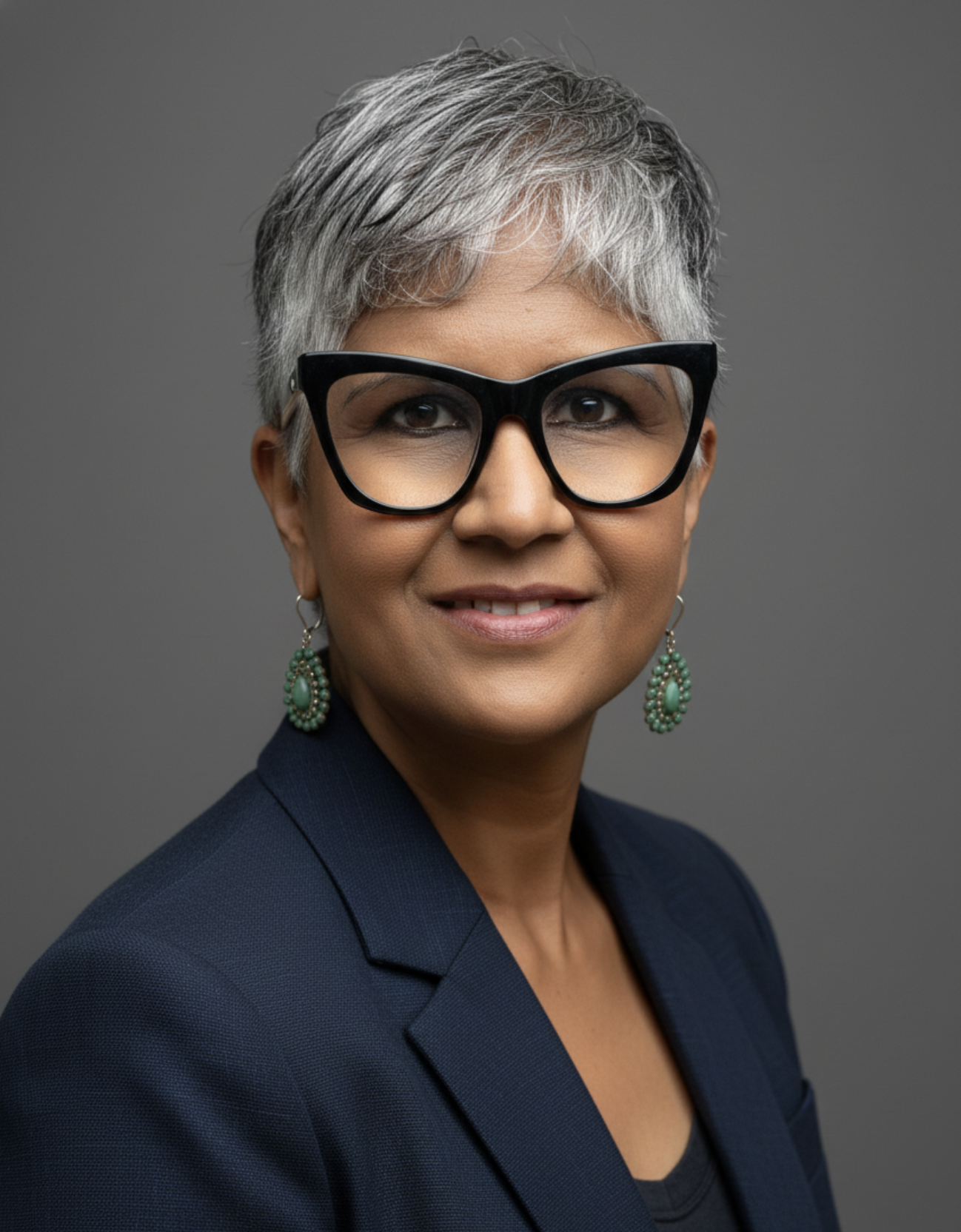 Smiling woman (Ateeka Anne Contee) with short gray hair, large black glasses, green earrings, and a light gray jacket outdoors