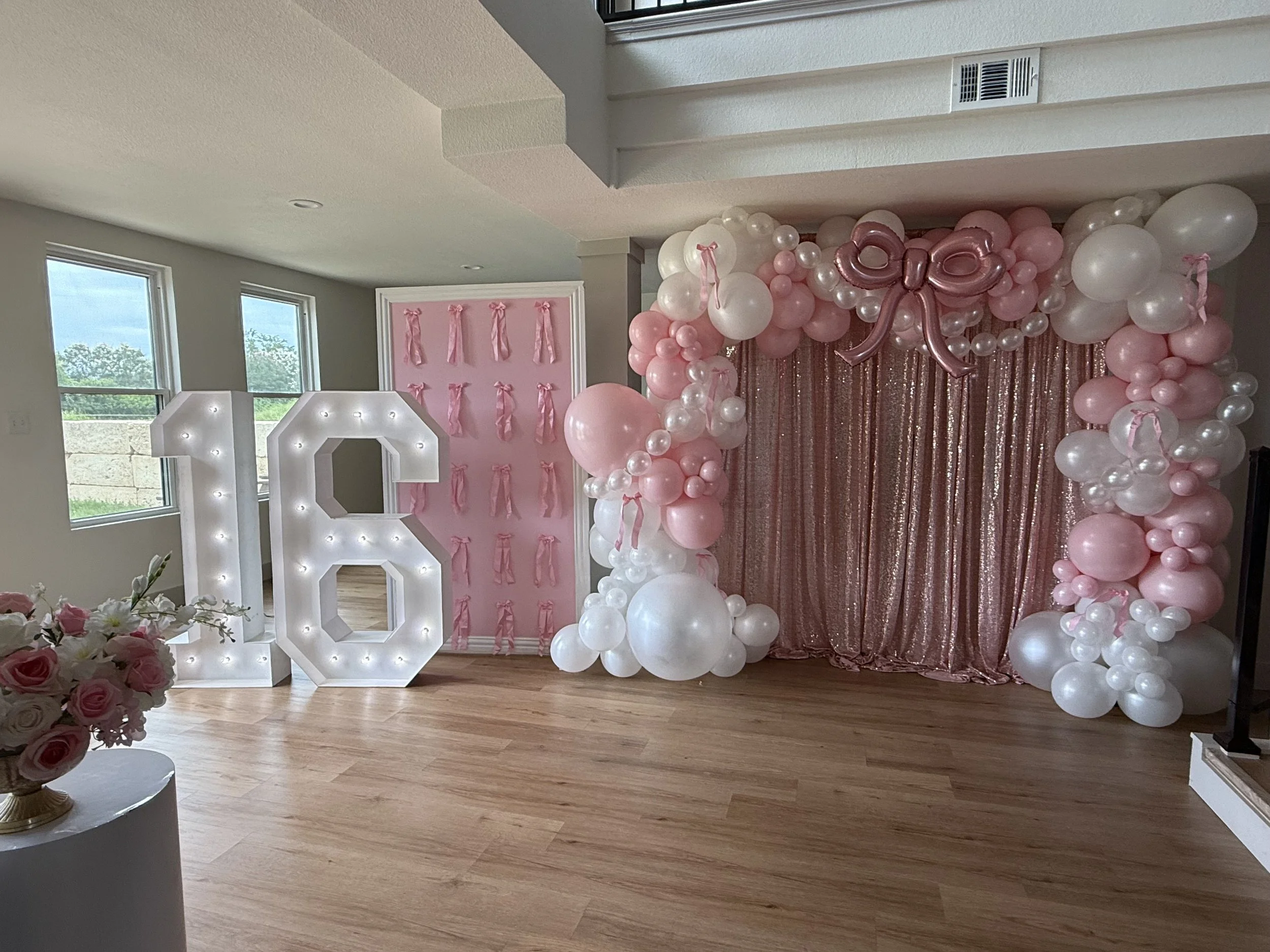 Decorated indoor space with large illuminated number six, pink and white balloon arch, pink sequin curtain backdrop, and pink ribbon wall decor.