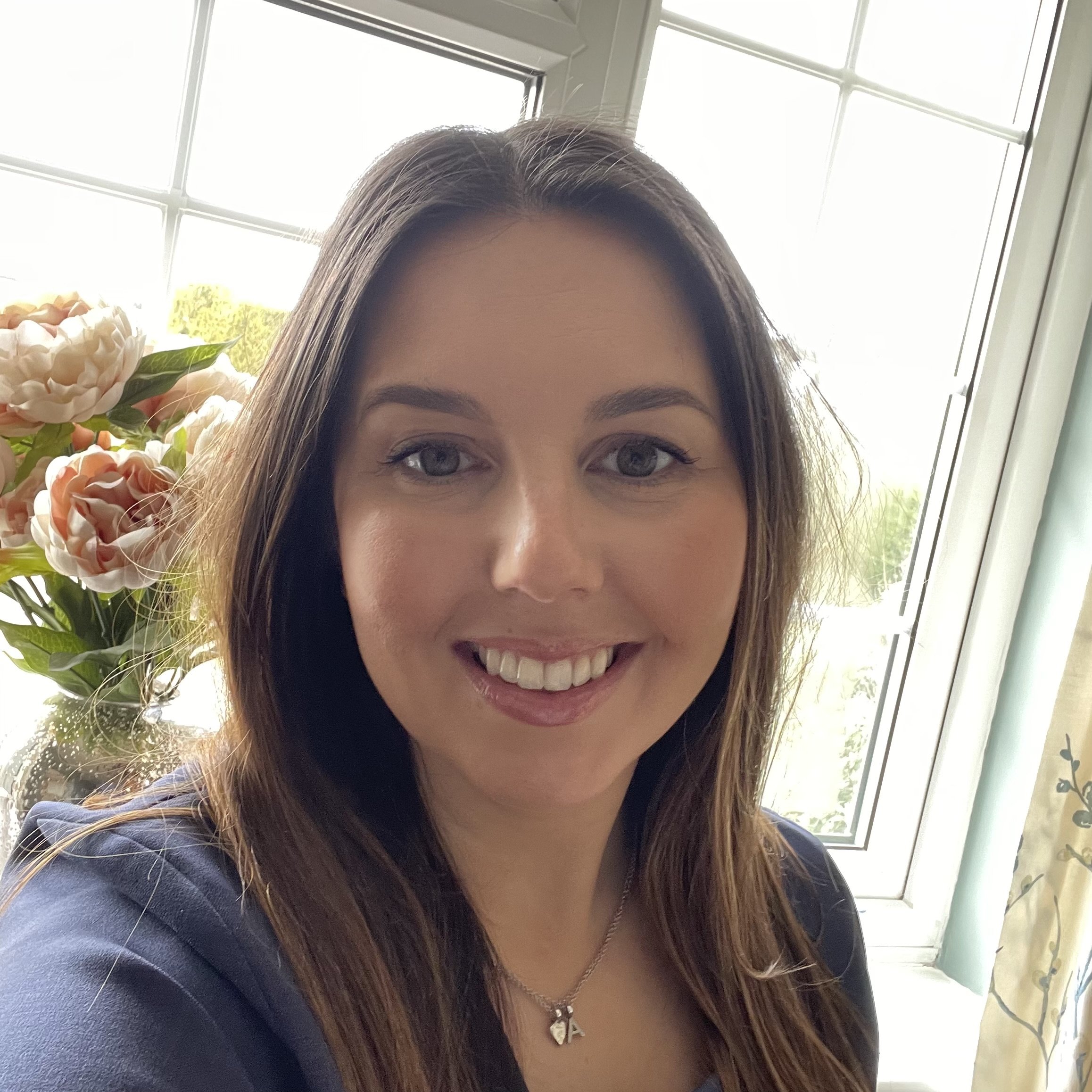 Smiling woman taking a selfie indoors with a window and flowers in the background.