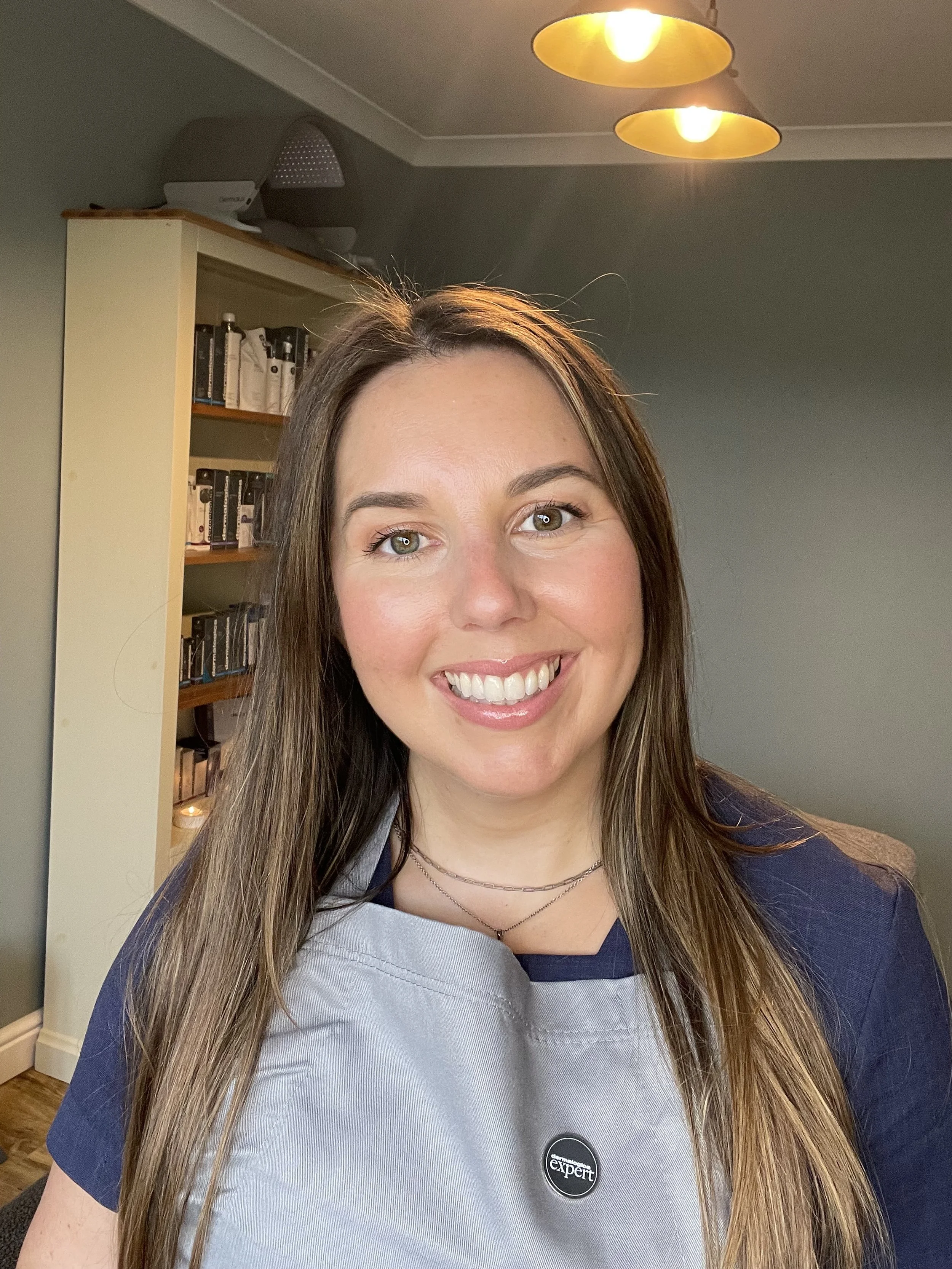 Smiling woman with long hair wearing an apron in a room with shelving and lights.