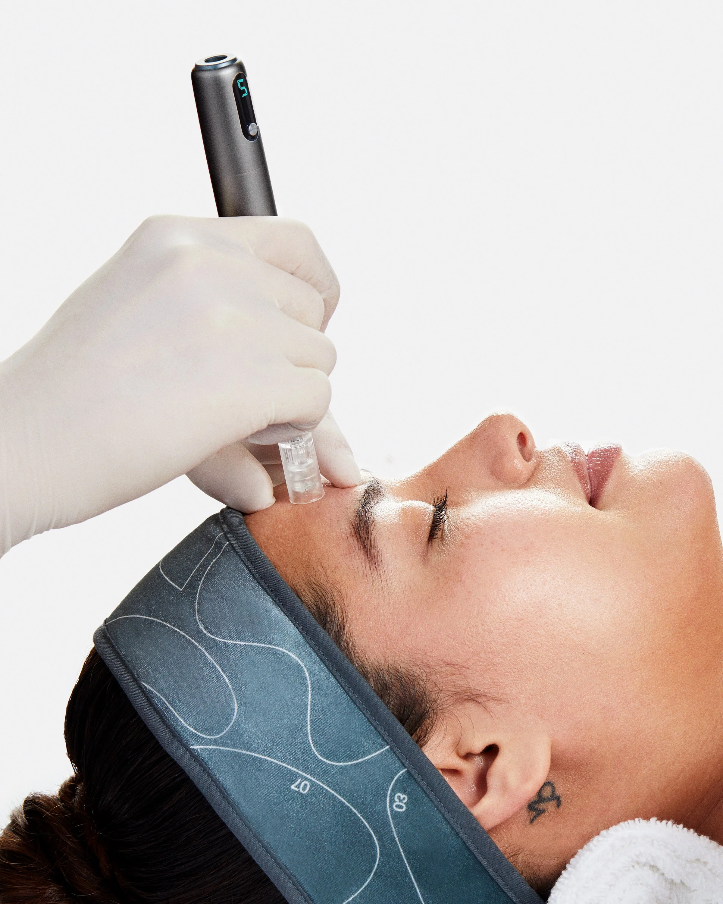 A person receiving a skincare treatment using a handheld device, with a headband on their forehead, applied by a gloved hand.
