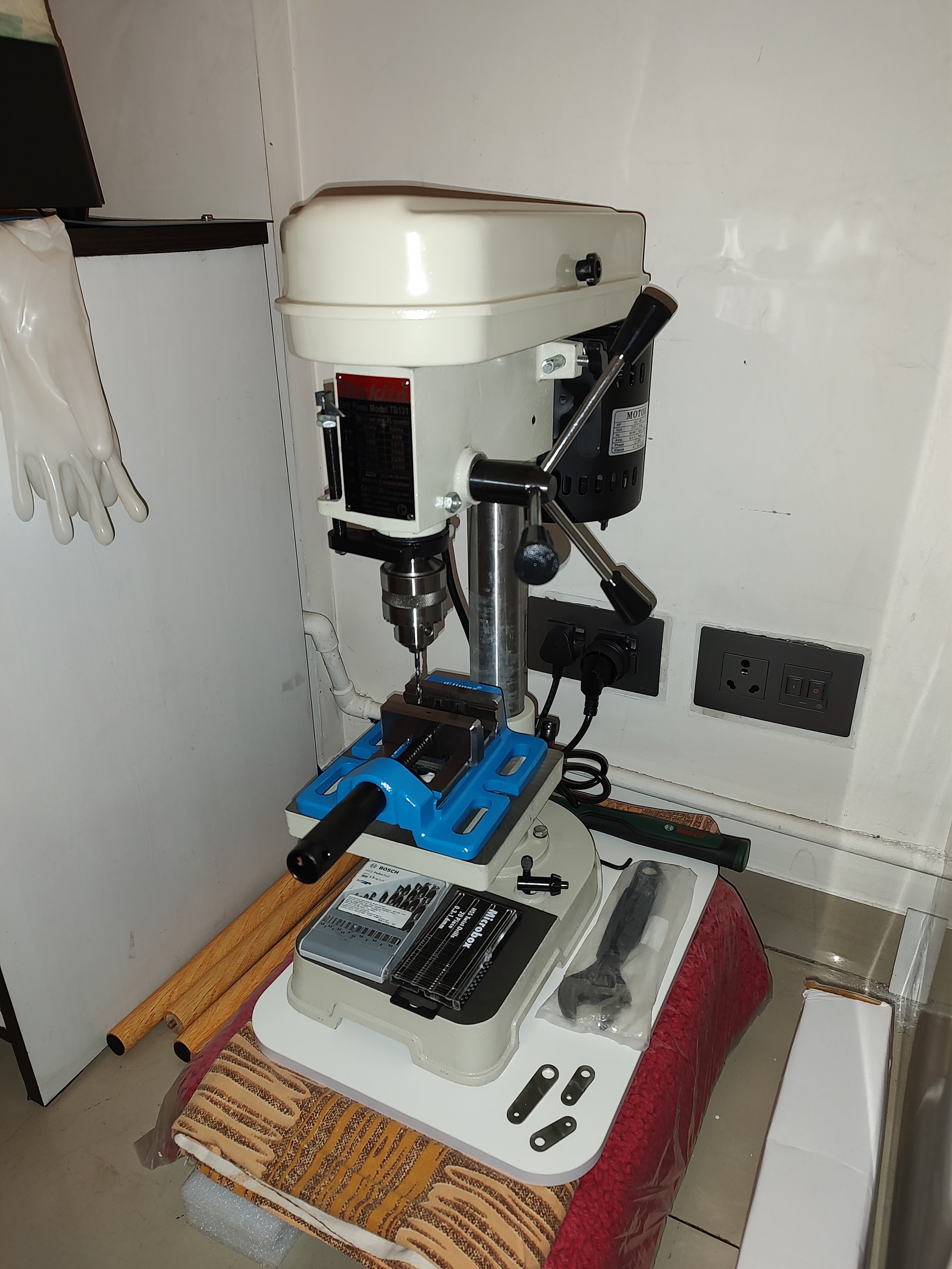 A mini drill press on a table with accessories nearby, set against a wall with electrical outlets.