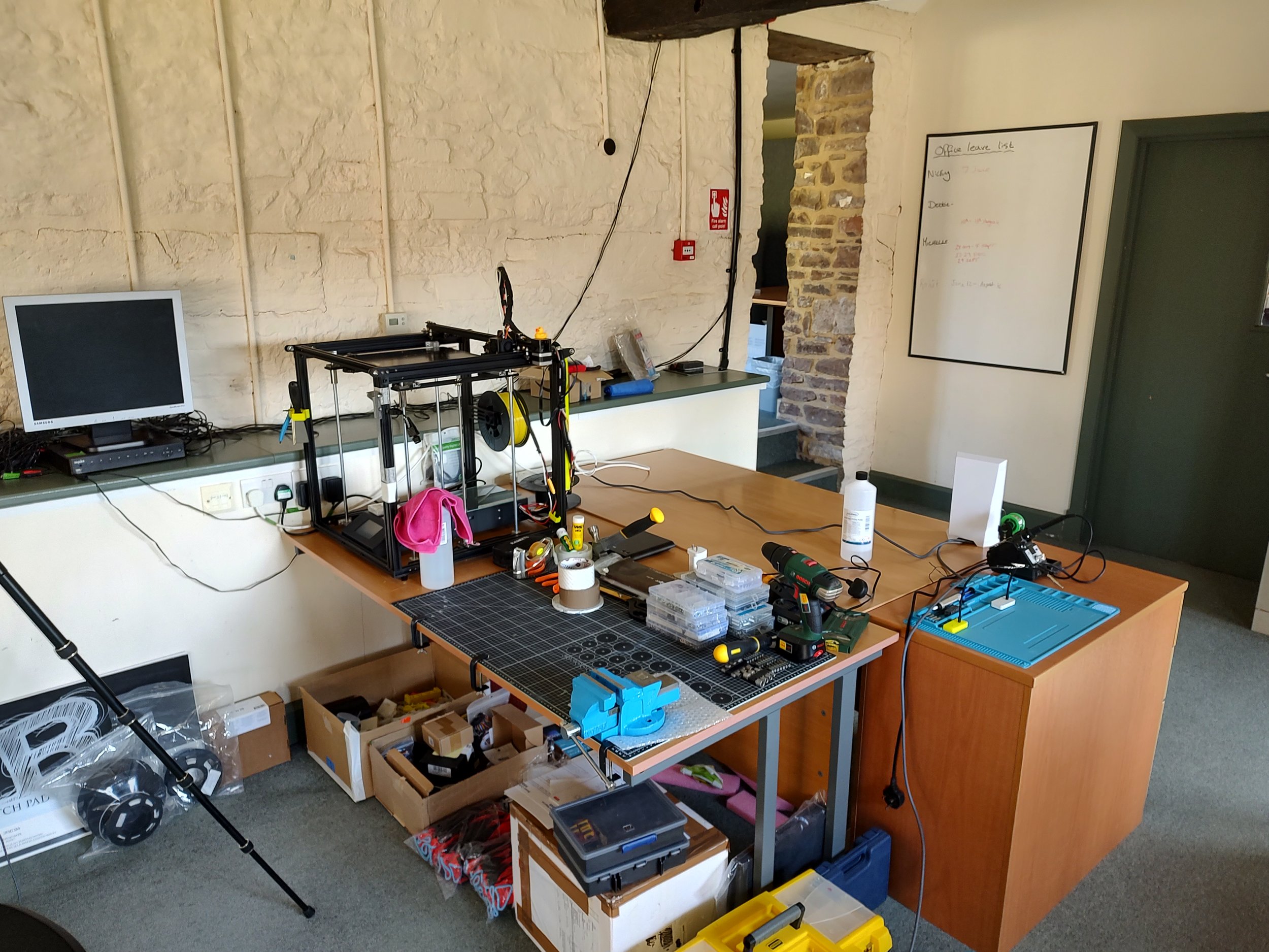 Workshop with 3D printer, tools, and electronics on a large wooden table, with a computer, whiteboard, and brick wall in the background.