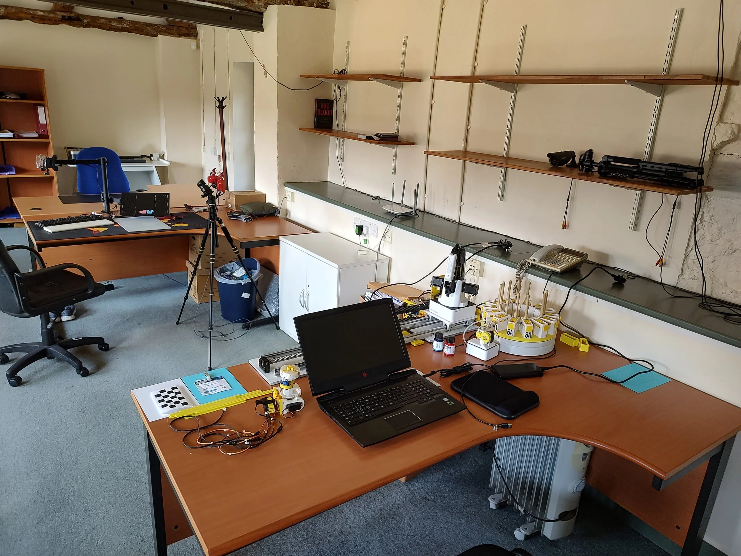 An office or laboratory workspace with desks, chairs, a laptop, scientific equipment, wires, and shelves.