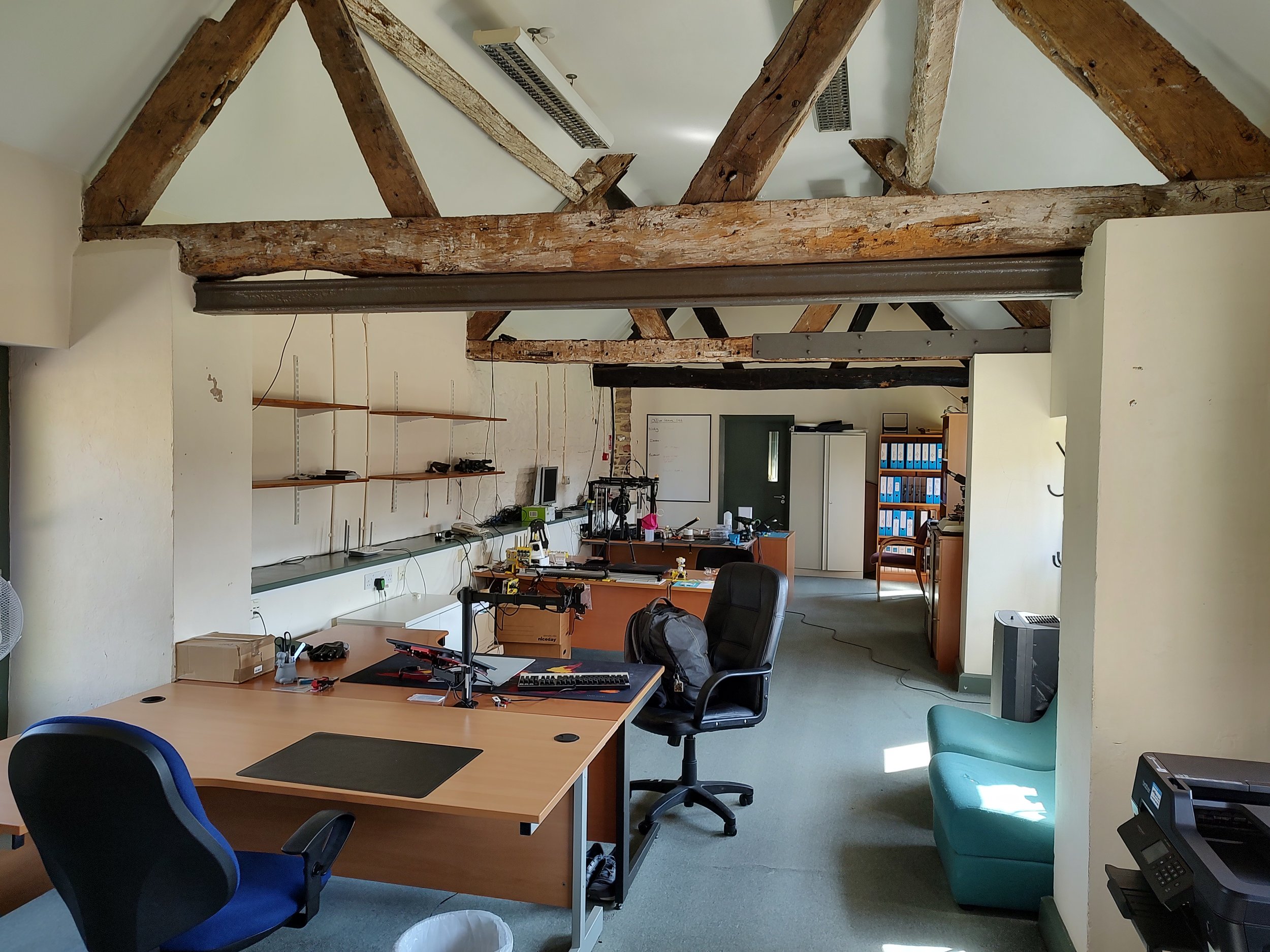 An office with wooden exposed beams on a sloped ceiling, desks, chairs, shelves, and filing cabinets.