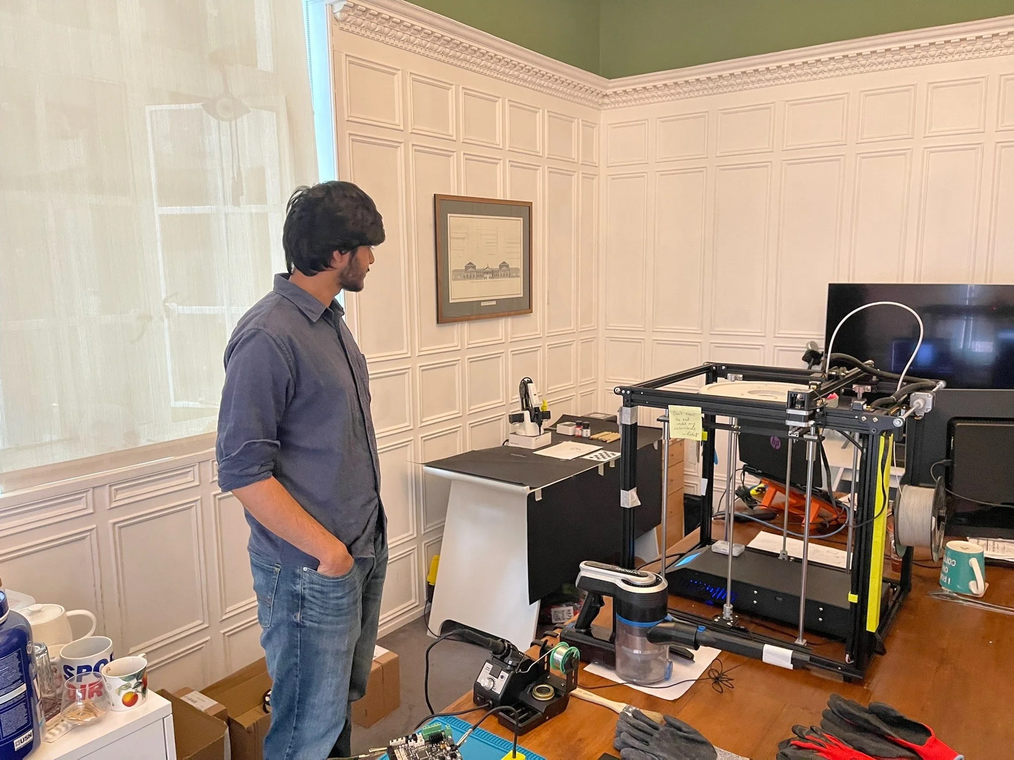 A man standing in a room with 3D printer equipment on a wooden table. The man is wearing a blue shirt with rolled-up sleeves and jeans, looking at the 3D printer. The room has white paneled walls, a framed picture, and various tools and supplies on t