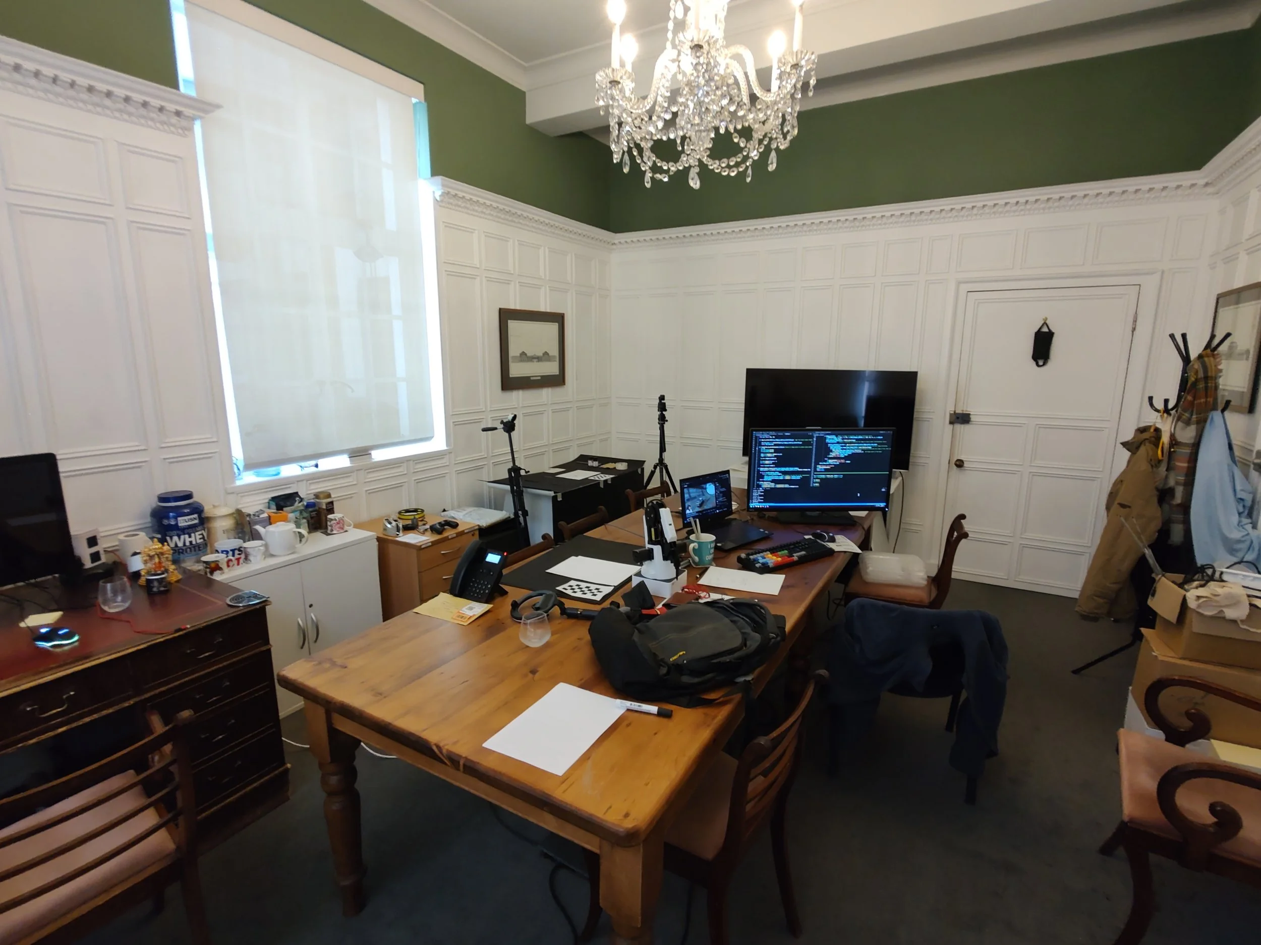 Office room with large wooden table, computer monitors, chairs, and various office supplies. Victorian-style white wall paneling, a crystal chandelier, and a window with white shades.