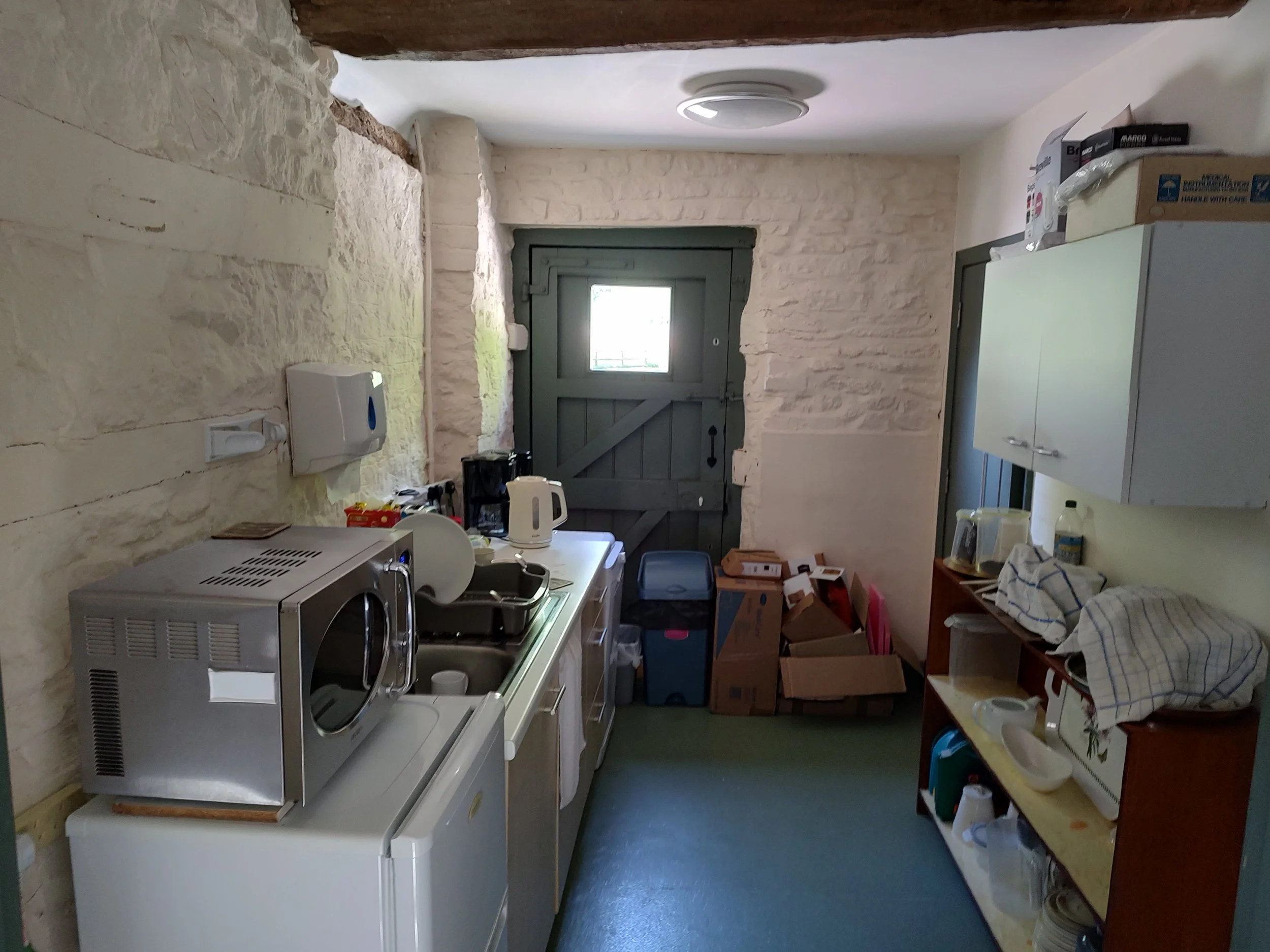 A small, cluttered kitchen with white stone walls, a windowed gray door at the end, and various household appliances and boxes.