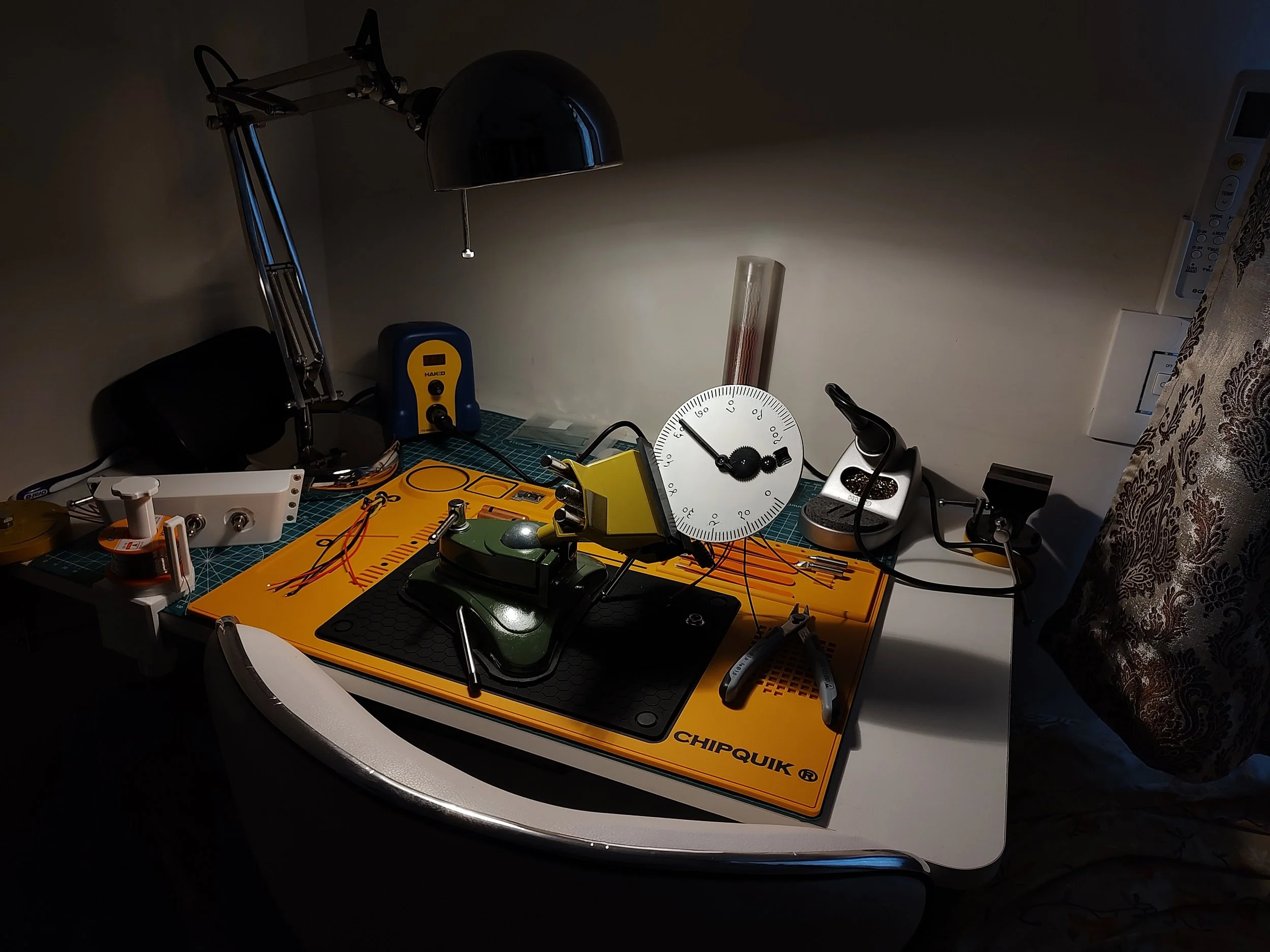 A workspace with an electronics workbench featuring various tools, components, and equipment, including a lamp, a yellow chipquik board, a soldering iron, a stopwatch, and wires on a cutting mat.