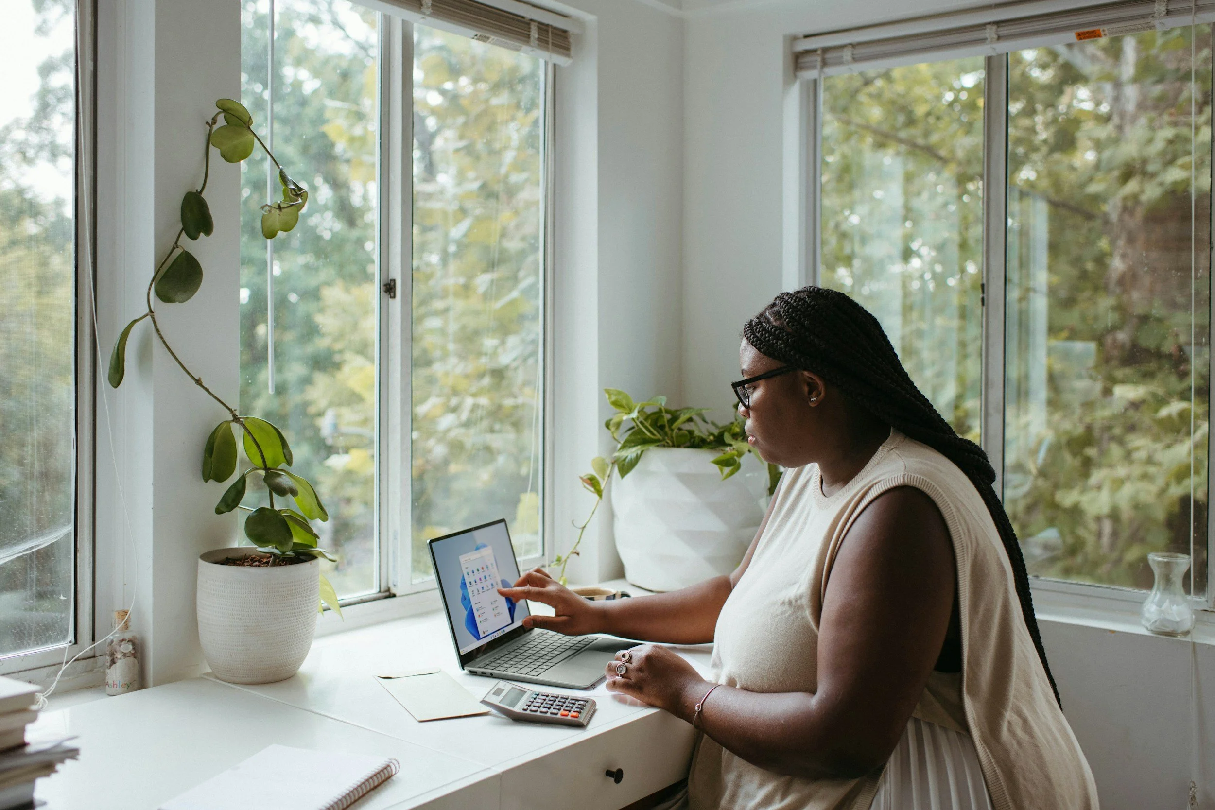 Woman with glasses using a laptop on a white desk in a bright room with large windows and indoor plants.