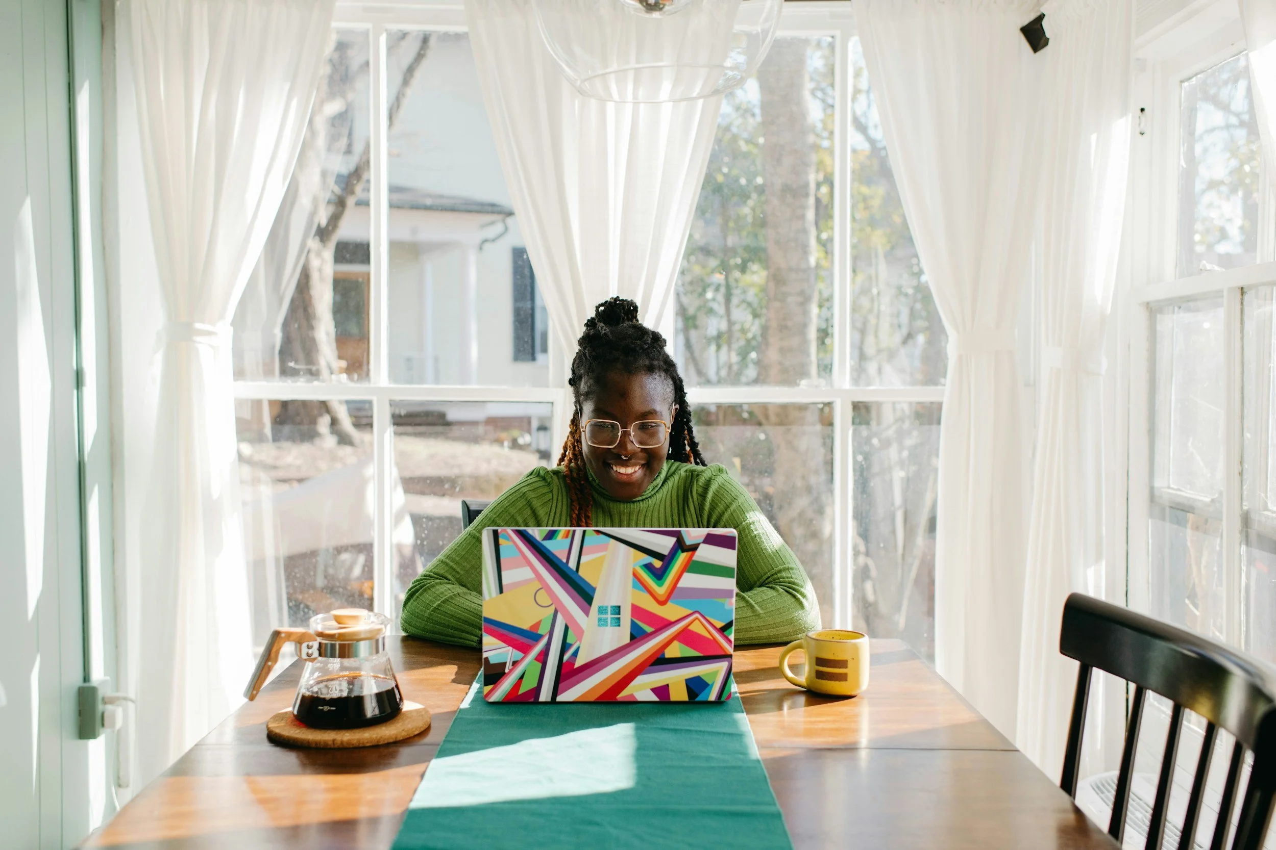 A woman with glasses and braids sitting at a table with a colorful laptop, a yellow mug, a glass coffee pot, and a wooden coaster in a bright room with large windows and white curtains.