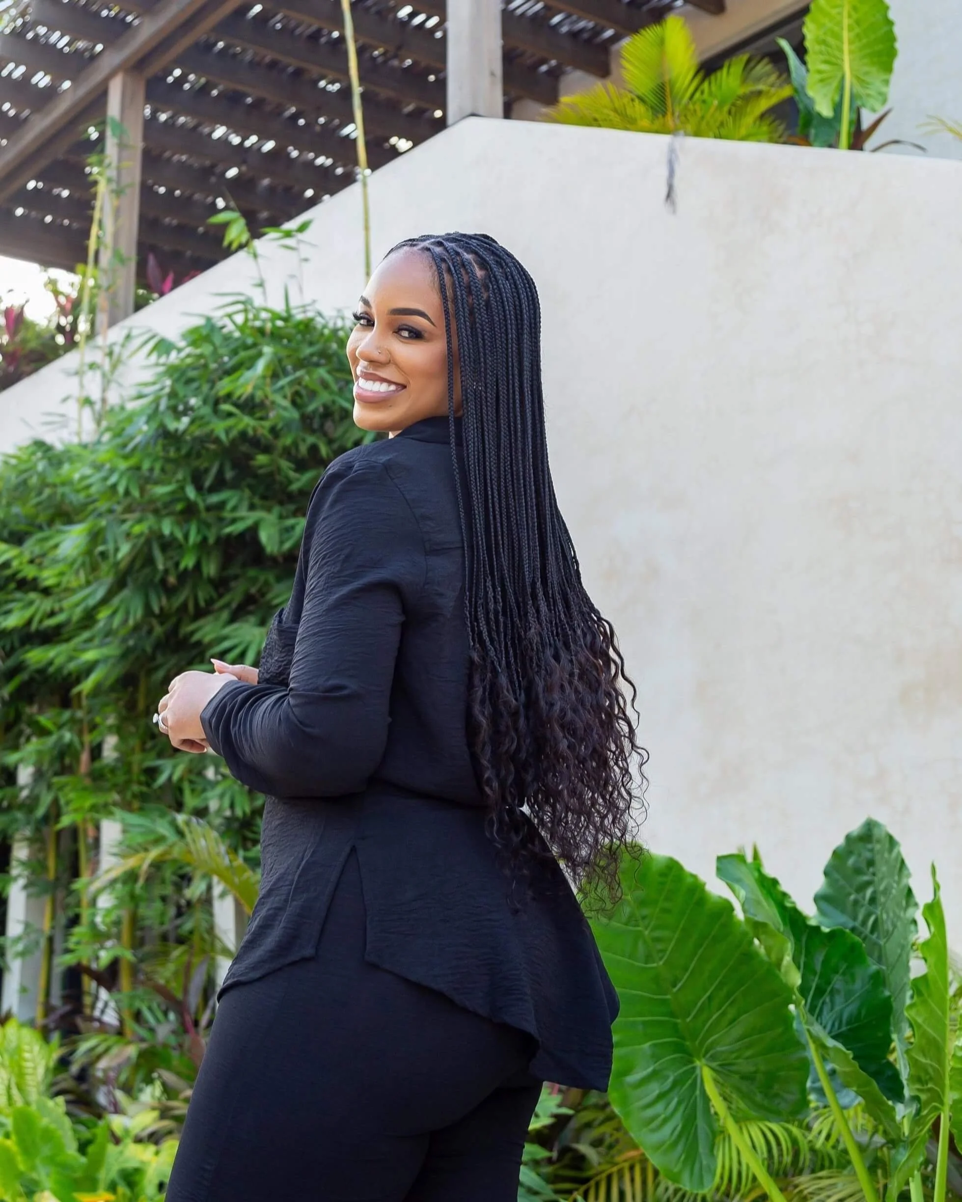 A smiling woman with long braids wearing a black jacket and black pants standing outdoors with green plants and a white wall in the background.