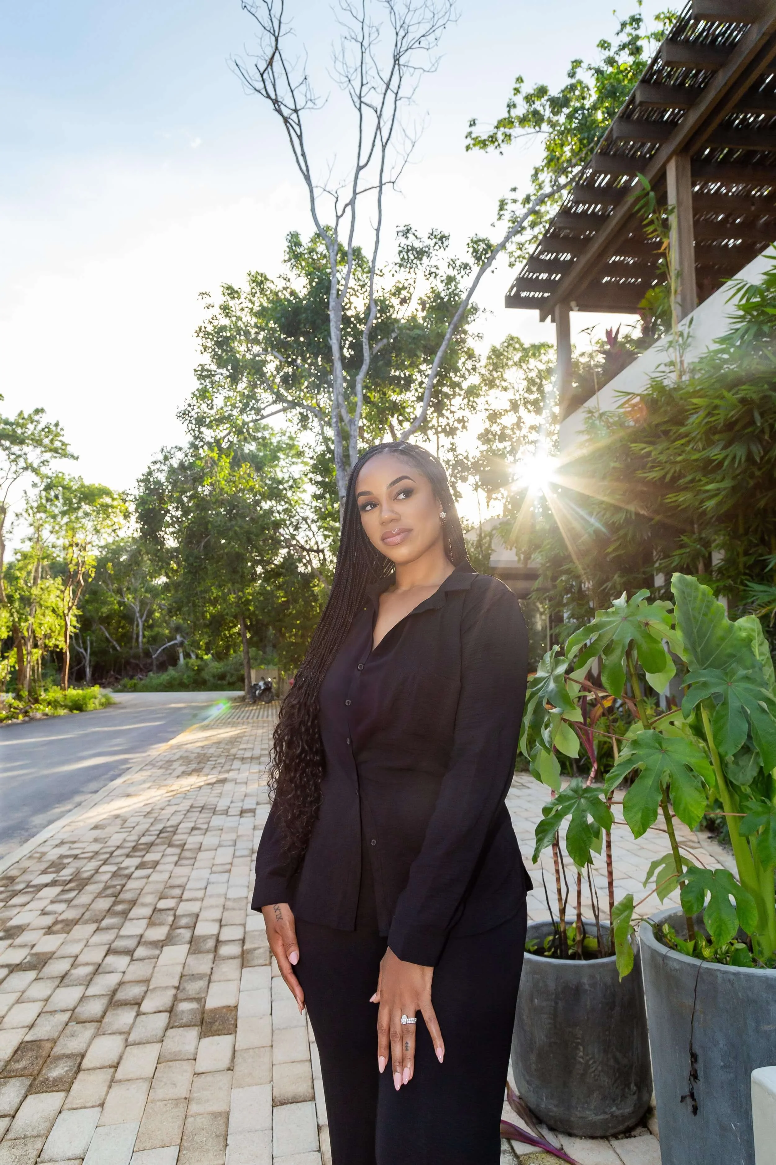 A young woman with long braided hair wearing a black blouse and black pants standing outdoors on a paved walkway, with greenery and potted plants around her, and the sun shining behind her creating a lens flare.
