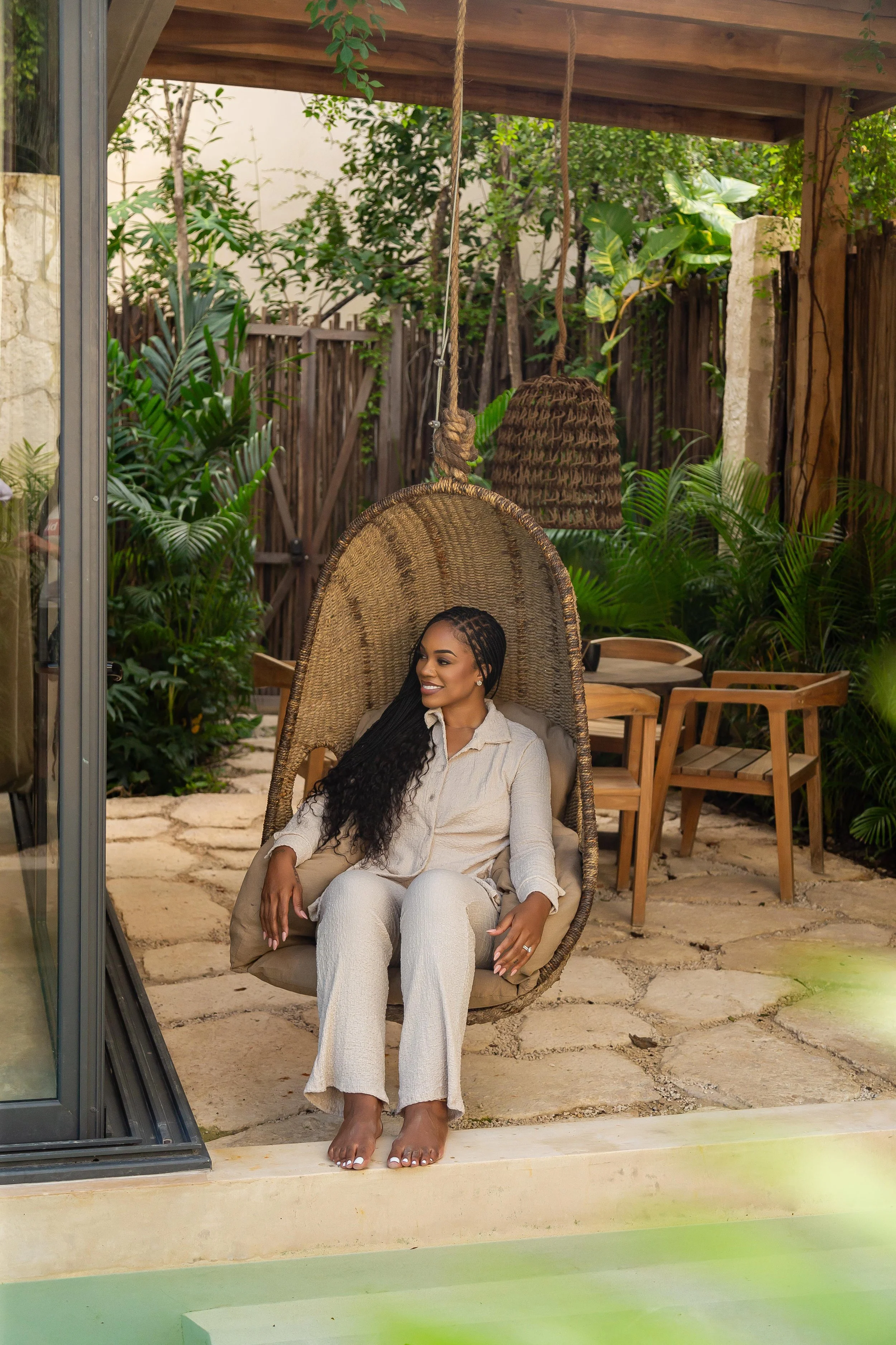 A woman with long braids sitting on a hanging wicker chair in a lush outdoor garden area with wooden furniture and greenery.