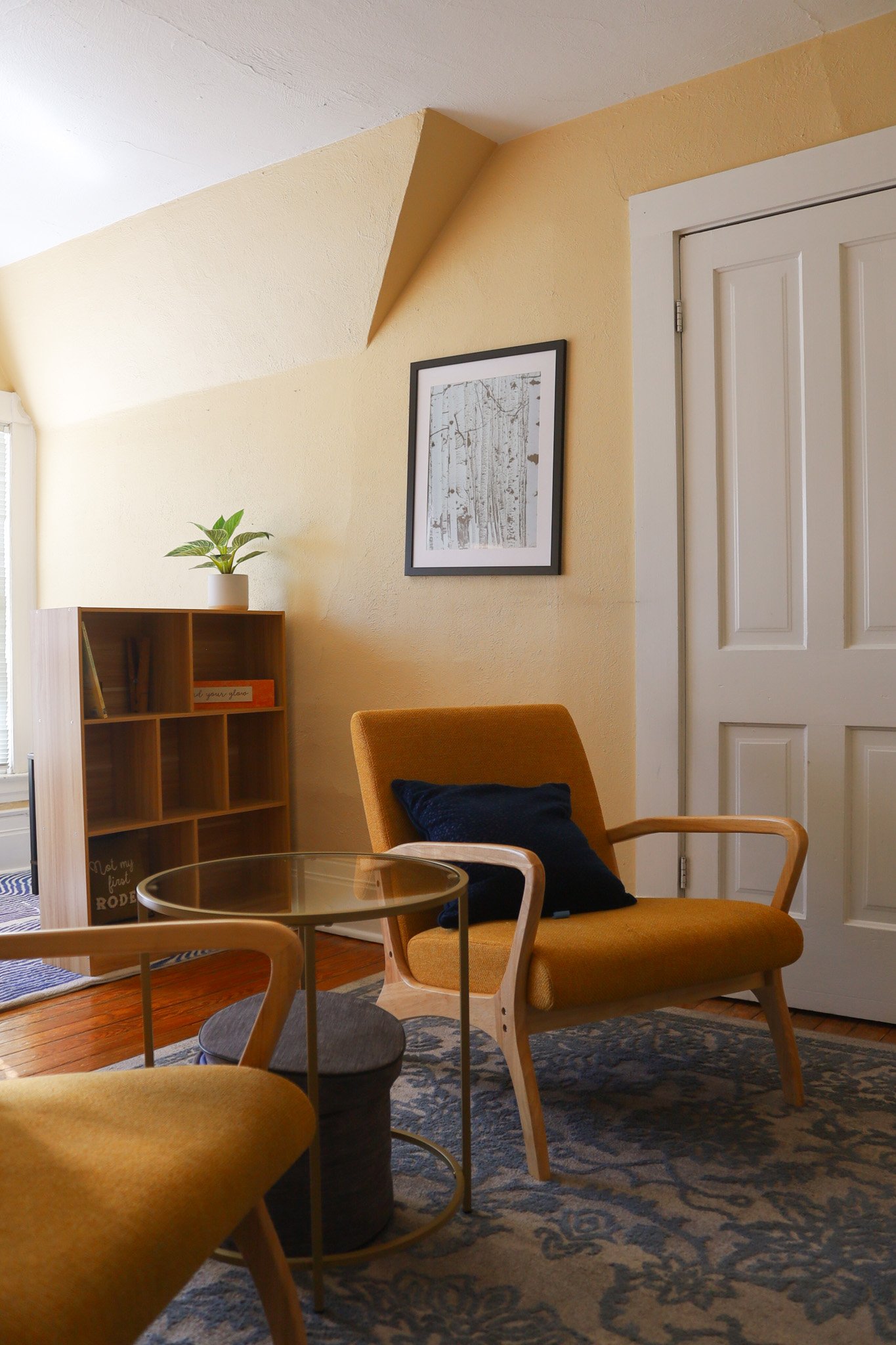 A cozy living room corner with a mustard yellow armchair, a black pillow, a small round glass-top side table, a wooden bookshelf with a potted plant, a framed black-and-white art piece, a patterned area rug, and a white door.
