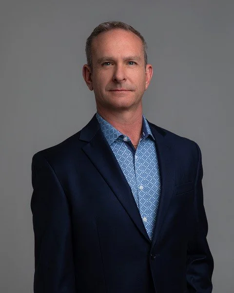 Man in a suit with a patterned shirt standing against a gray background.
