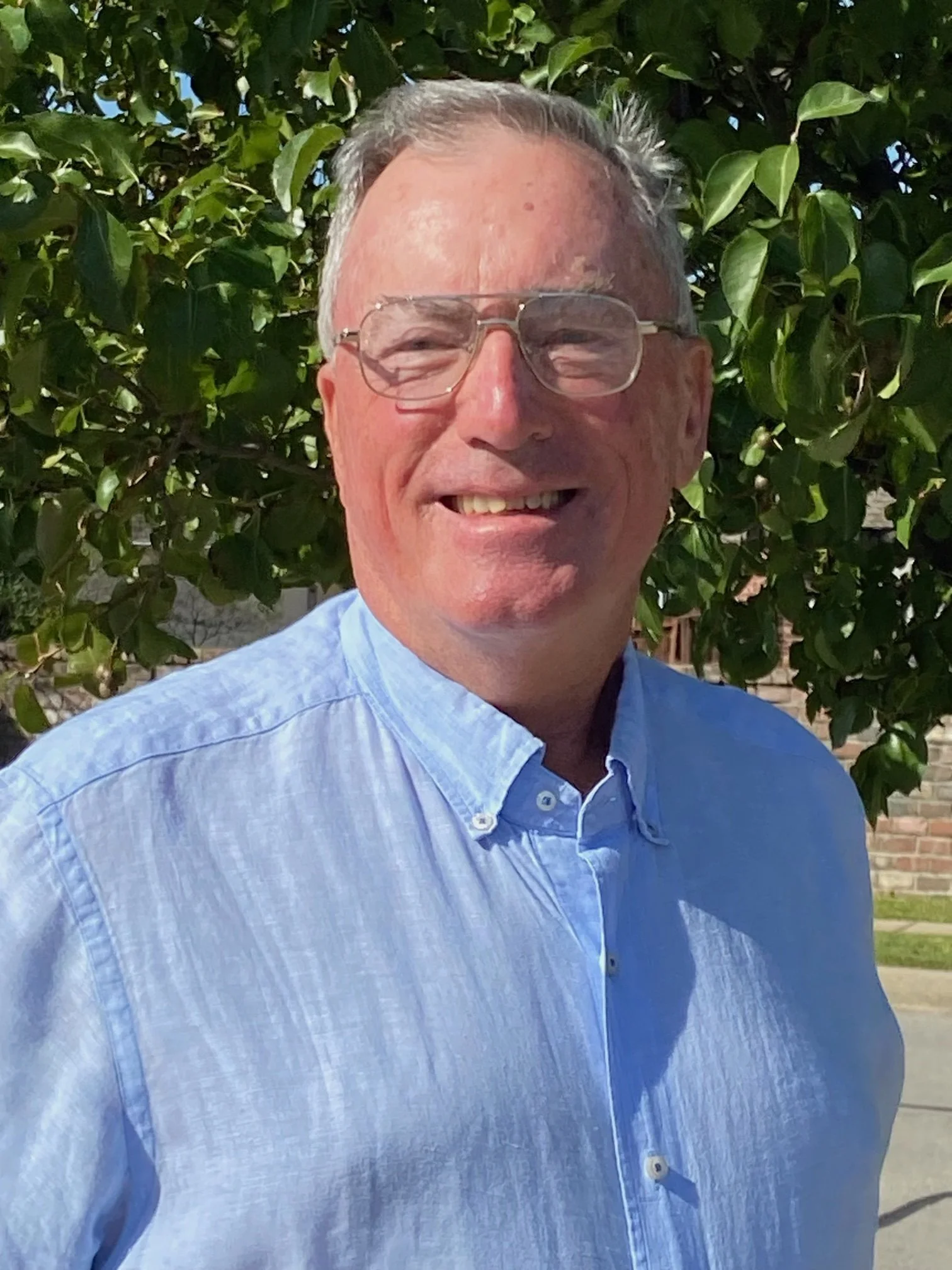 Smiling person wearing glasses and a blue shirt standing outdoors with trees in the background.