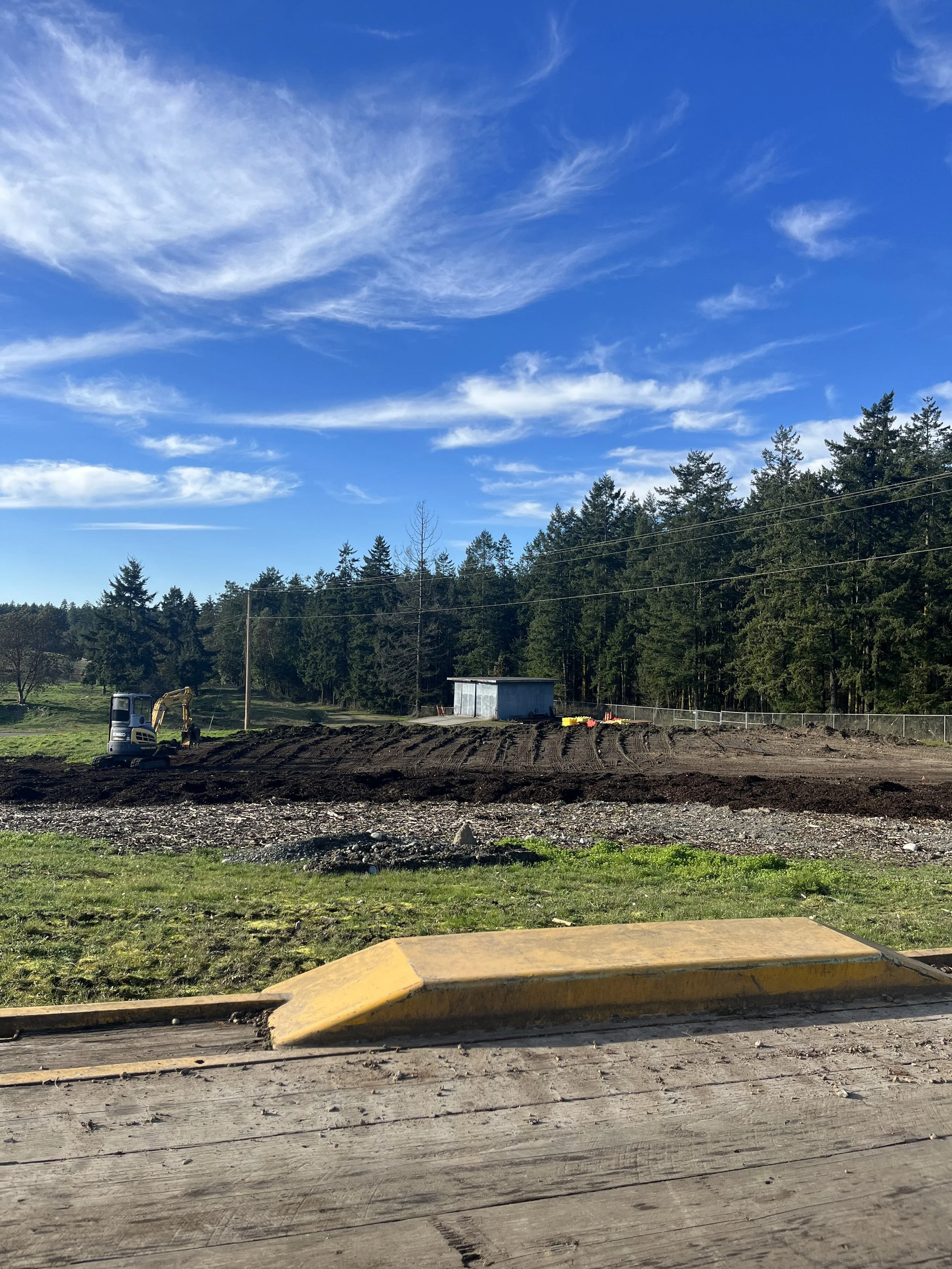 Construction site with a small excavator, cleared land, and trees in the background under a blue sky with clouds.