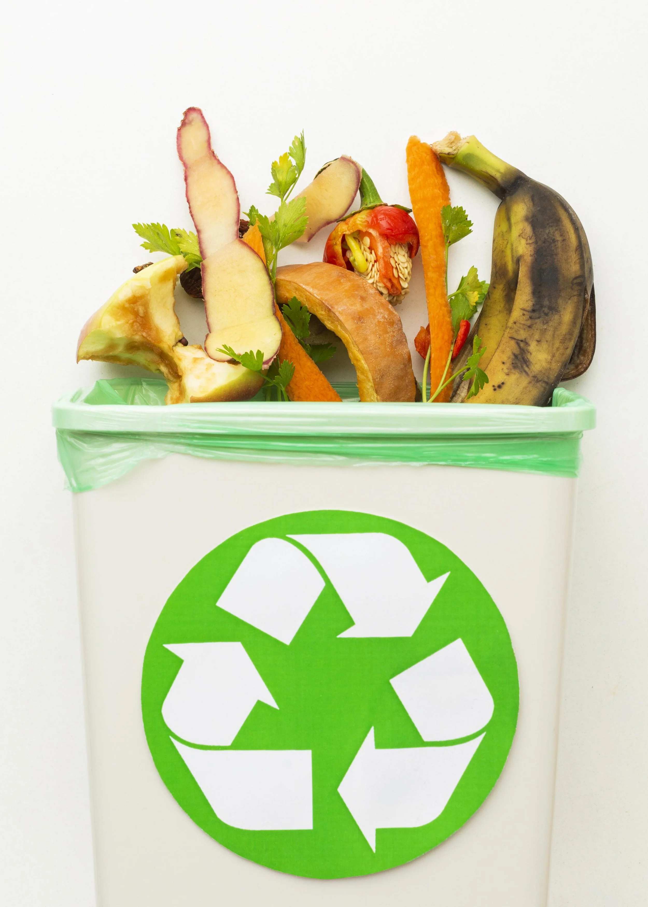 A white recycling bin with a green recycling symbol on the front, filled with various vegetable and fruit peels such as banana, apple, orange, and other produce scraps.