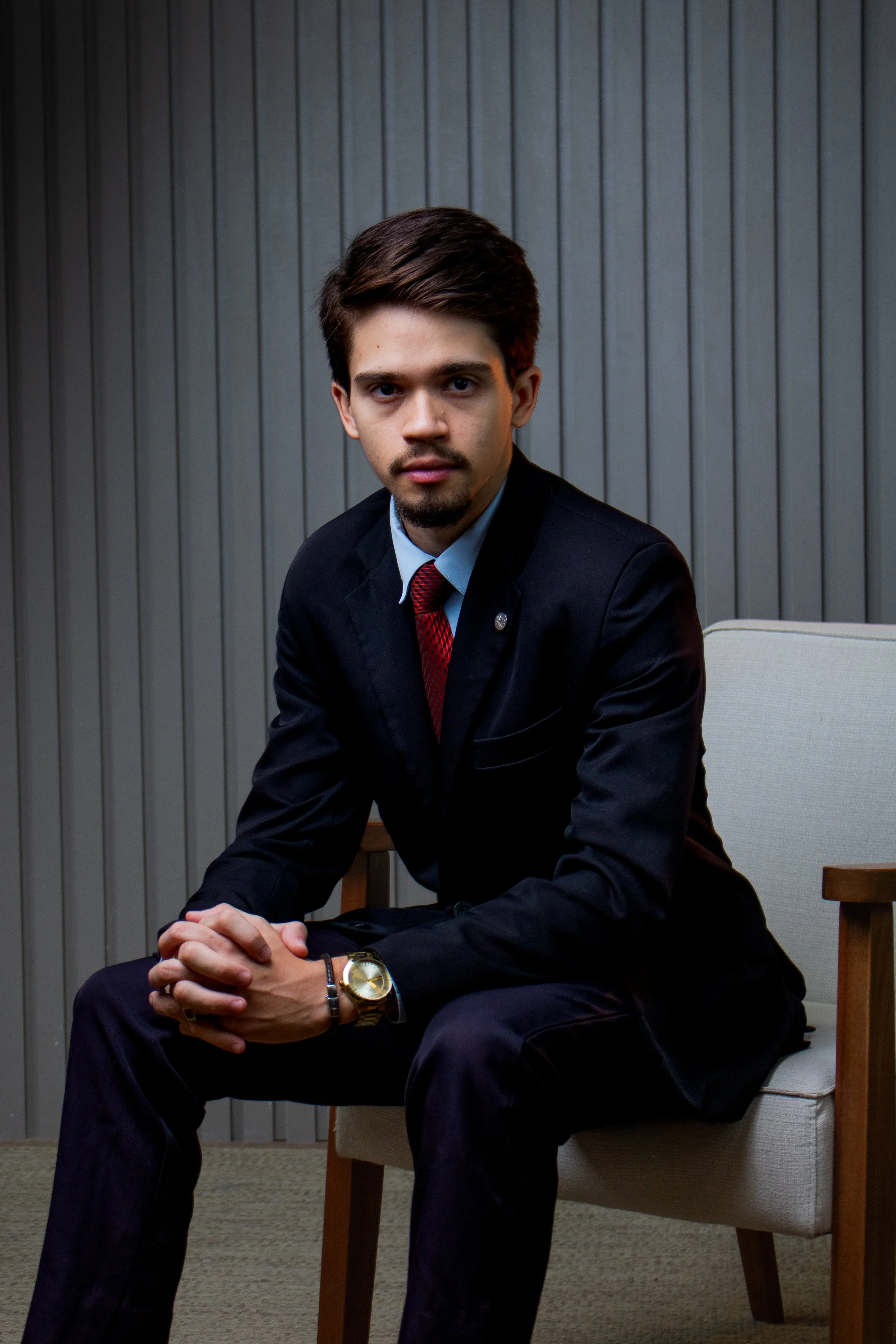 Homem jovem de terno preto, camisa azul claro, gravata vermelha, com cabelo castanho, barba por fazer, sentado em uma cadeira de tecido cinza com braços de madeira, diante de uma parede de painéis de madeira cinza vertical, olhando para a câmera com expressão séria.