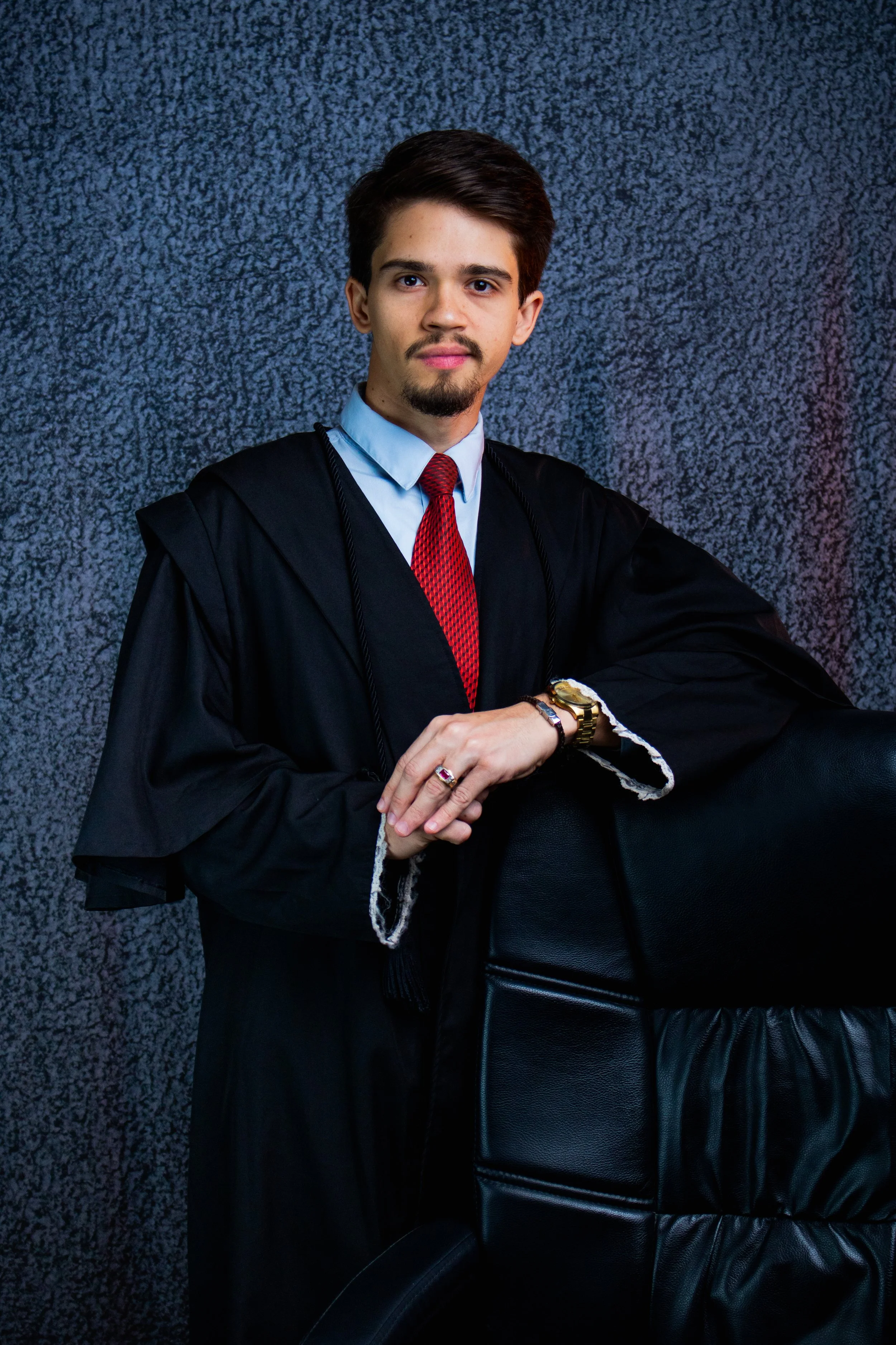 Homem jovem em traje académico, usando beca preta, camisa azul e gravata vermelha, posando ao lado de uma cadeira de escritório de couro preto.