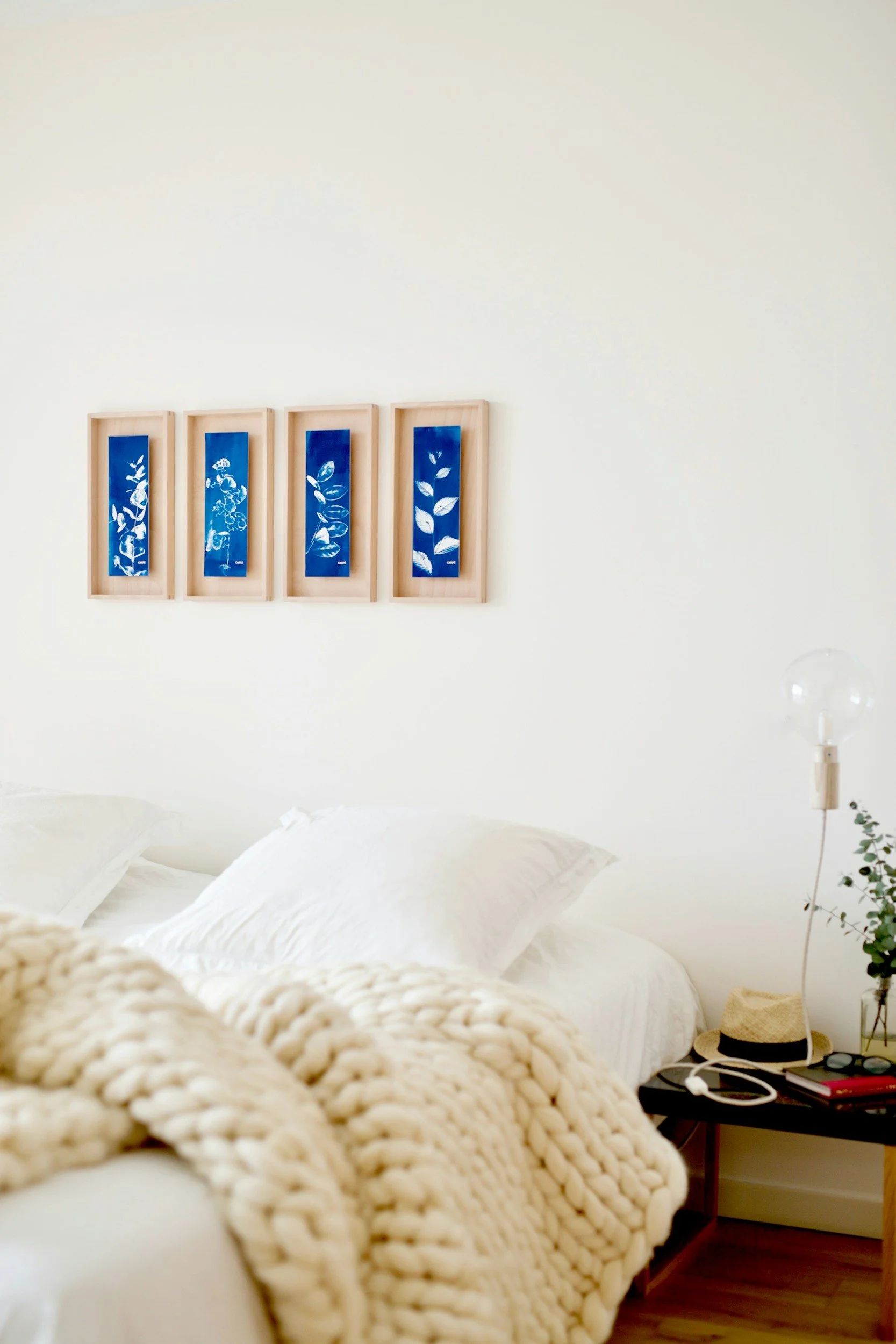A cozy bedroom features white bedding and pillows, with a chunky knit blanket in the foreground. On the wall above, four framed blue botanical prints are aligned horizontally. A side table holds a glass lamp, a straw hat, and some small plants.