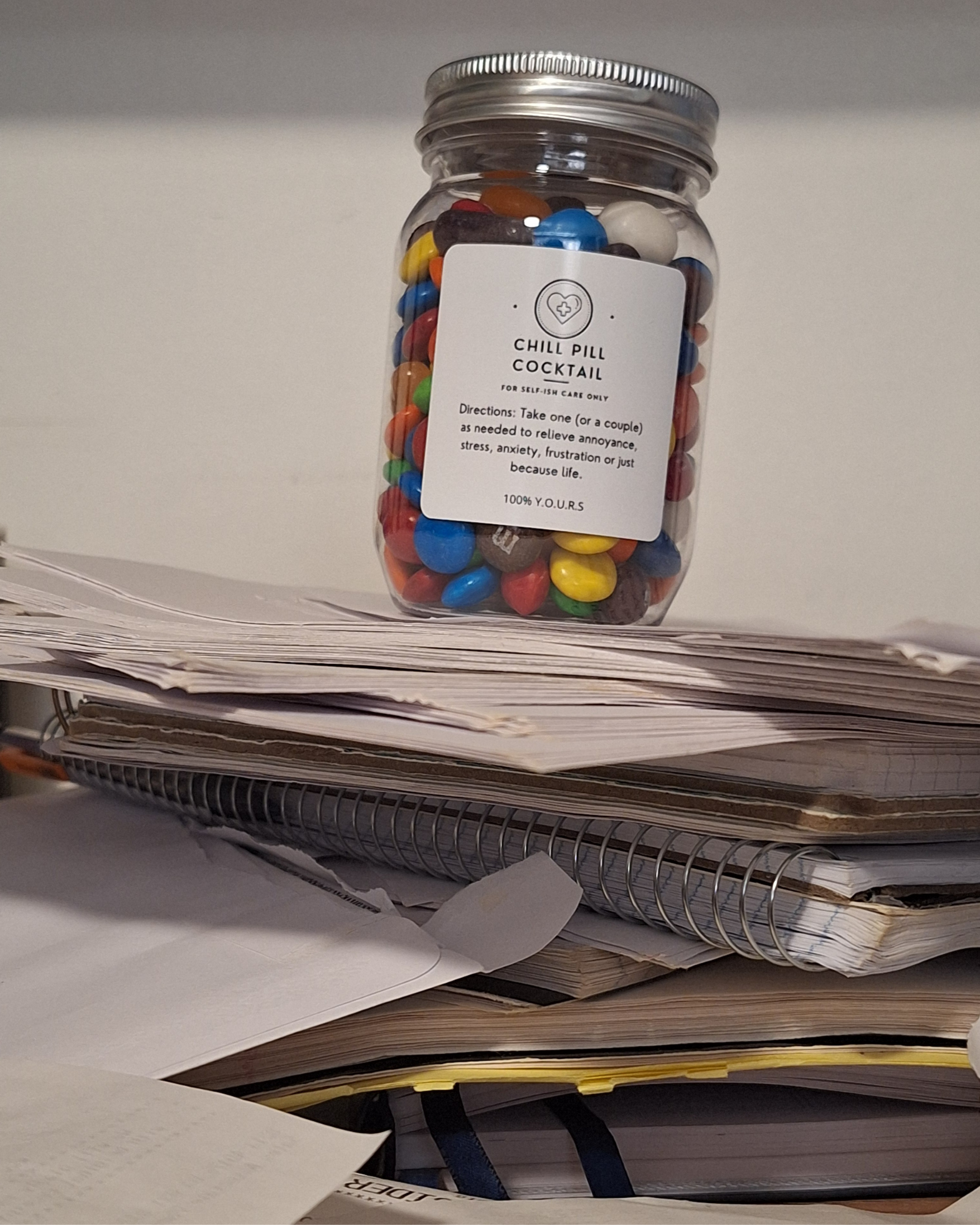 A jar labeled 'Chill Pill Cocktail' containing colorful candies, sitting on a cluttered desk with stacks of papers and notebooks.