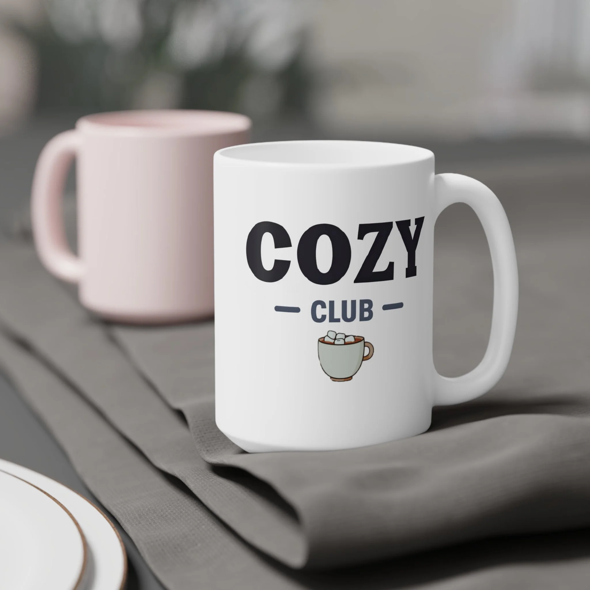 A white coffee mug with the text "COZY CLUB" and a small illustration of a mug filled with marshmallows on it, placed on a grey cloth, with a blurred pink mug in the background.