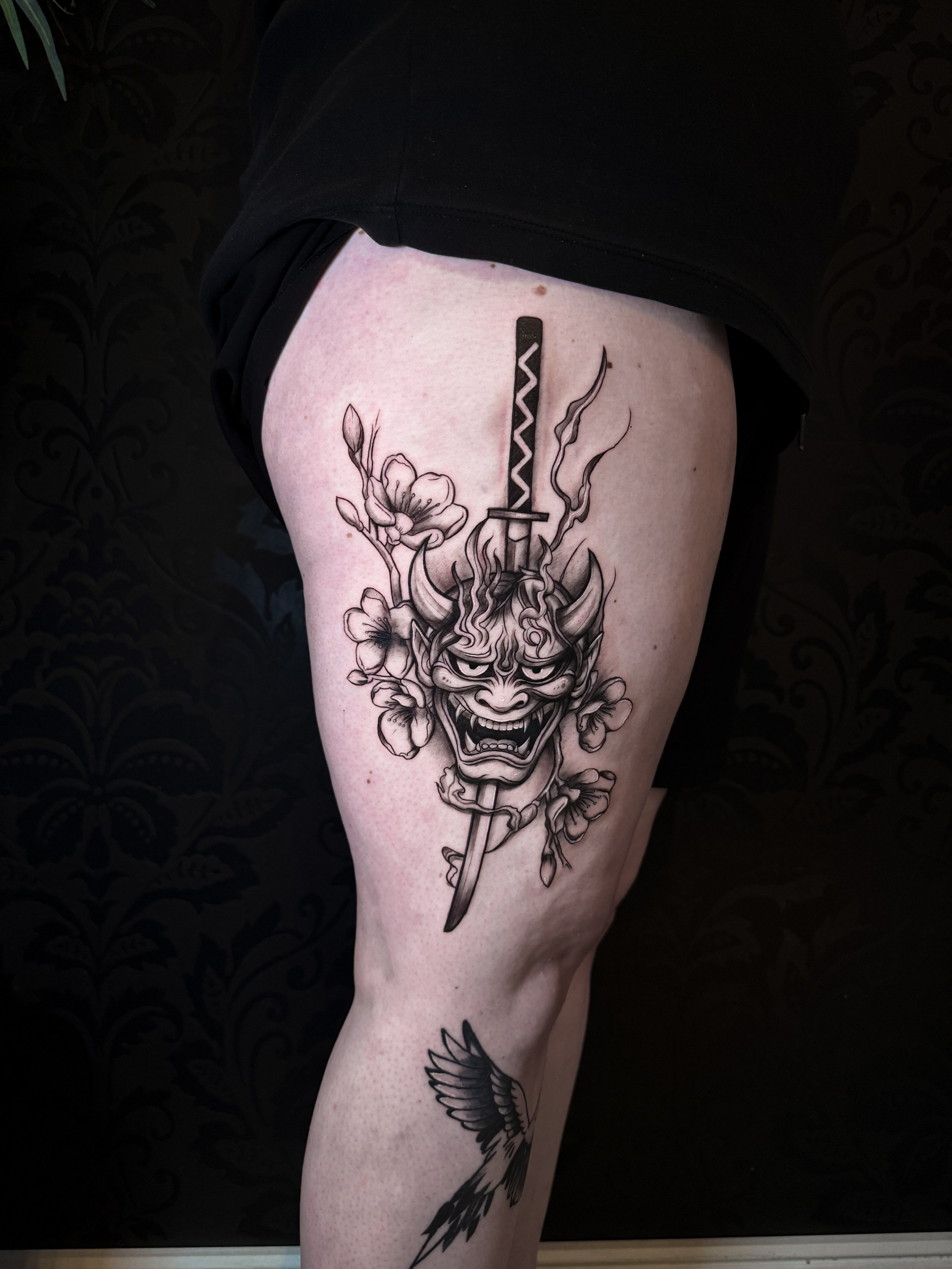 Hannya-Maske mit Katana-Schwert, floralen Elementen und Rauch im Black-and-Grey-Stil, großflächig auf den äußeren Oberschenkel tätowiert.