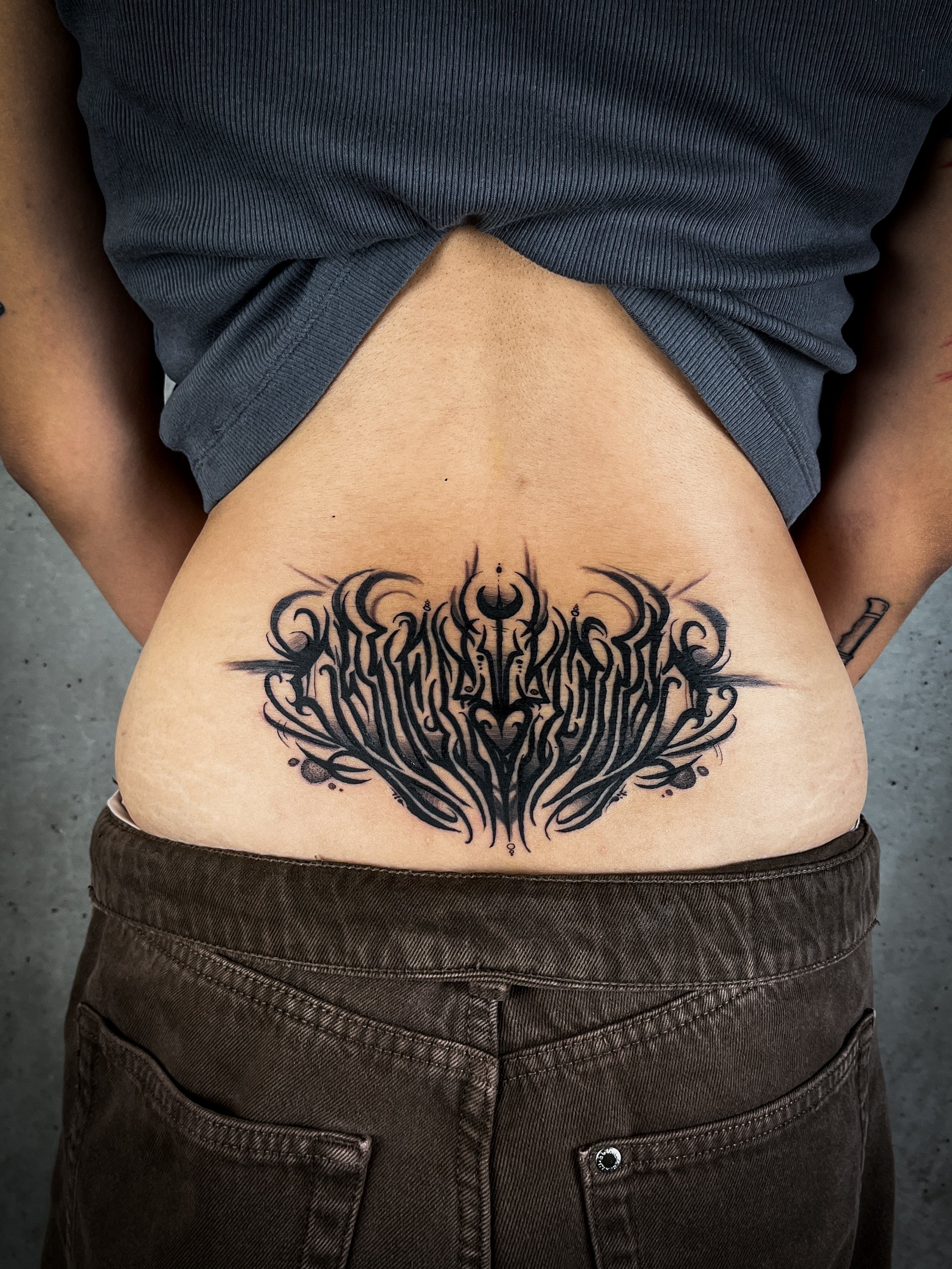 Blackwork-Lettering auf dem unteren Rücken, symmetrisch und ornamental aufgebaut, gestochen im klaren, dunklen Schriftstil.