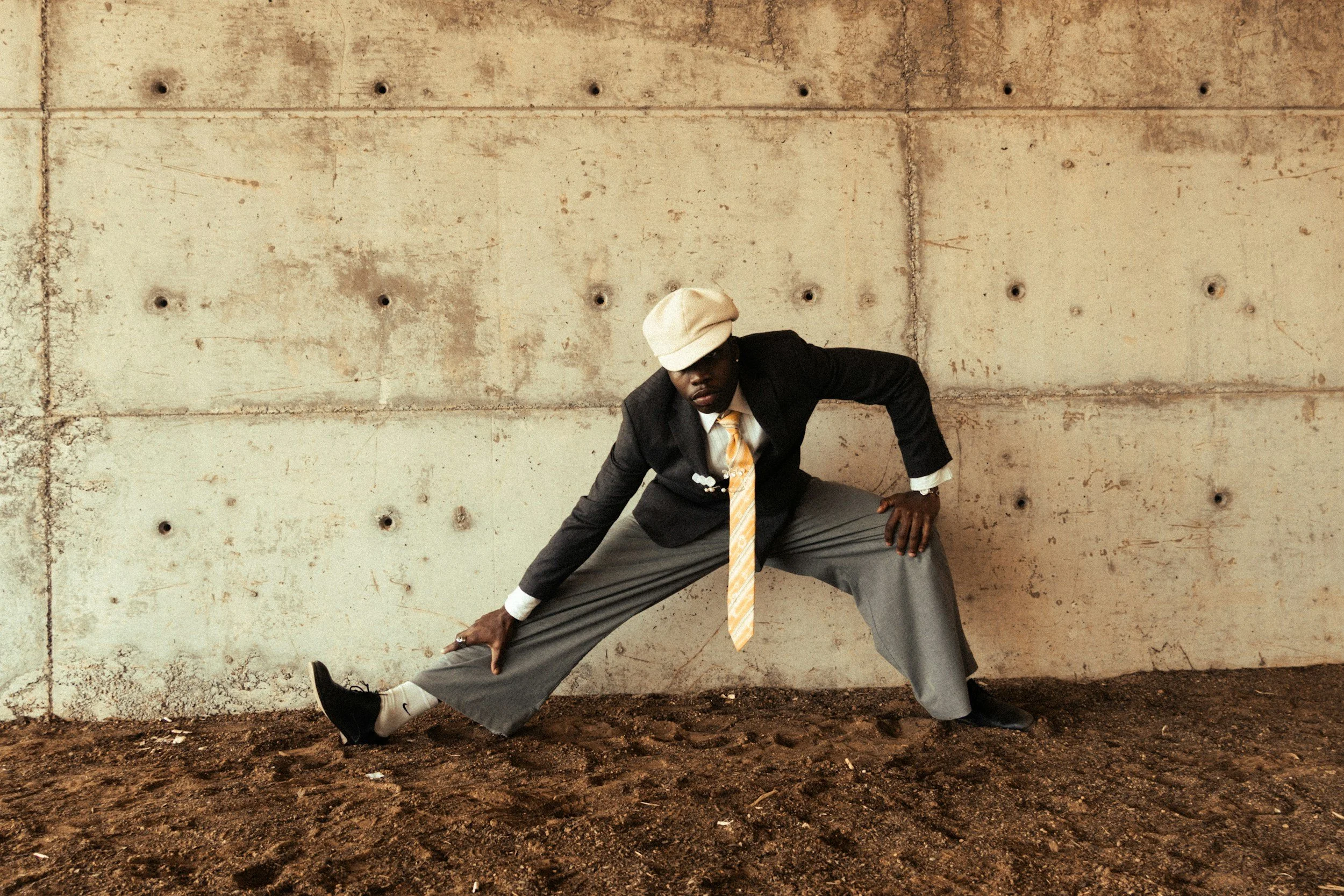 A man dressed in a black suit, yellow tie, and beige hat is posing in front of a plain, textured concrete wall. He is stretching his legs apart in a wide stance, with one hand touching the ground and the other reaching to the side.
