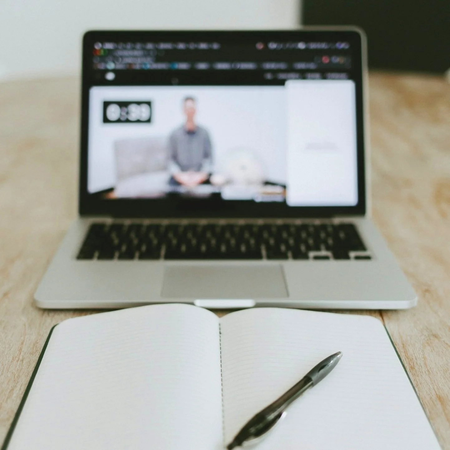 Open laptop displaying a blurred video call or webinar with an open lined notebook and pen in the foreground on a wooden desk.