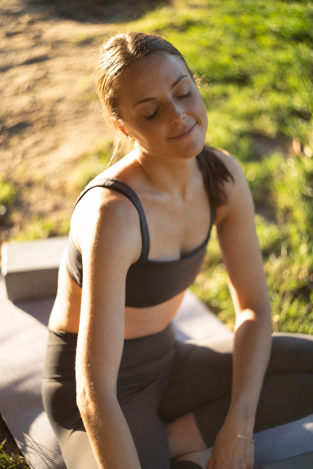 A woman doing yoga outdoors on a mat in natural sunlight with eyes closed and a peaceful expression, surrounded by grass and dirt.