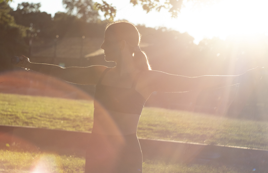 A woman practicing yoga outdoors at sunset with arms extended sideways, backlit by the sun.