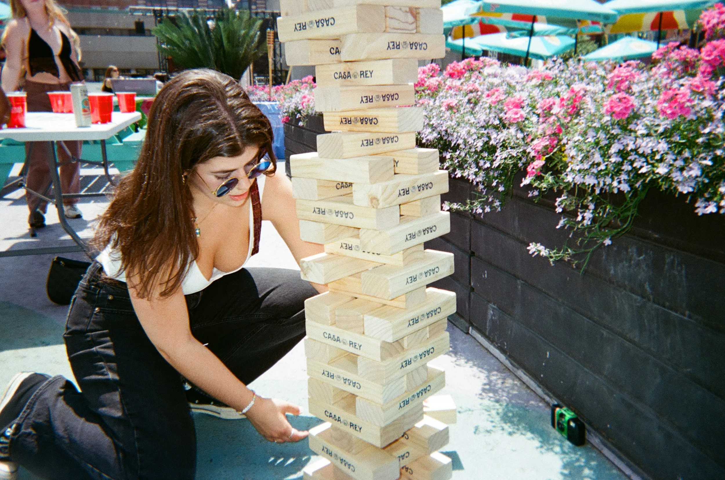 A woman wearing sunglasses and casual clothing plays a giant Jenga game outdoors on a sunny day, with pink and purple flowers in the background.