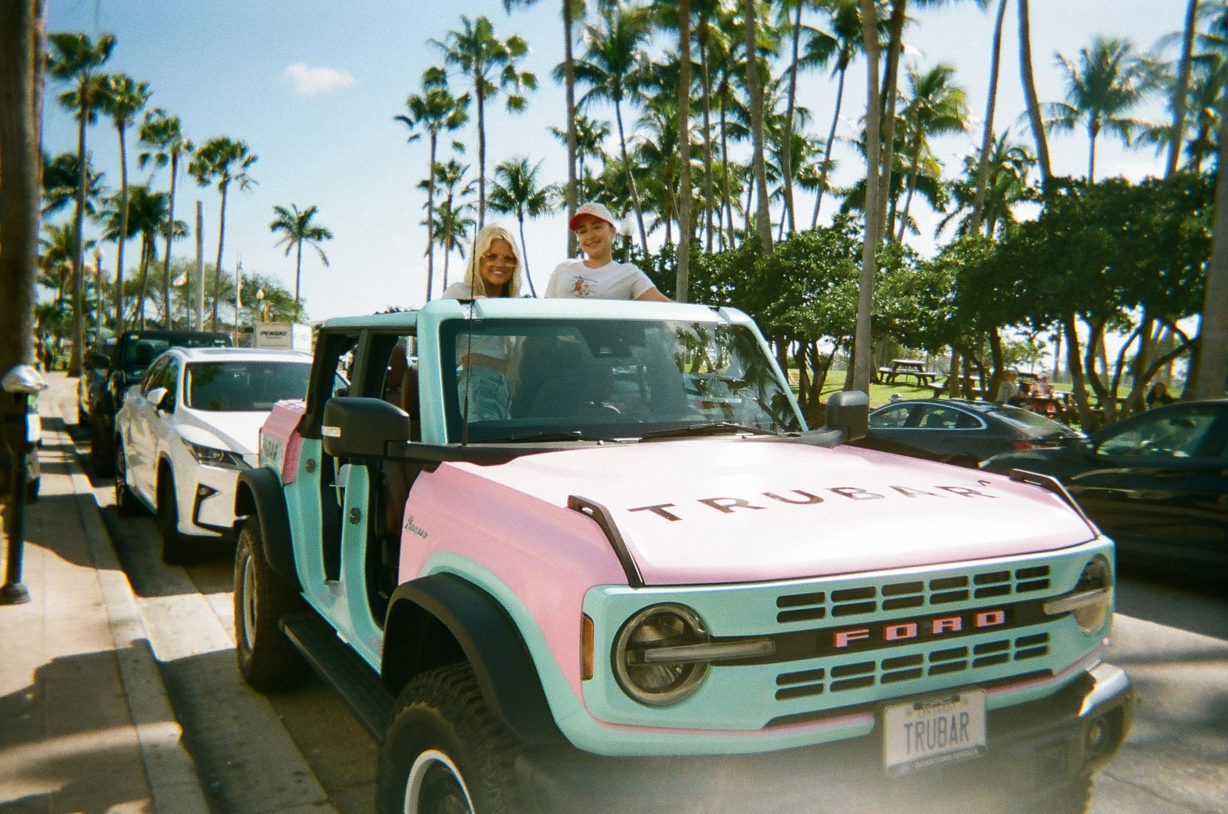 A pastel-colored Ford vehicle with the word Trubar on the front, parked in a line of cars at a location with many tall palm trees, with two women smiling and standing inside the vehicle on a sunny day.