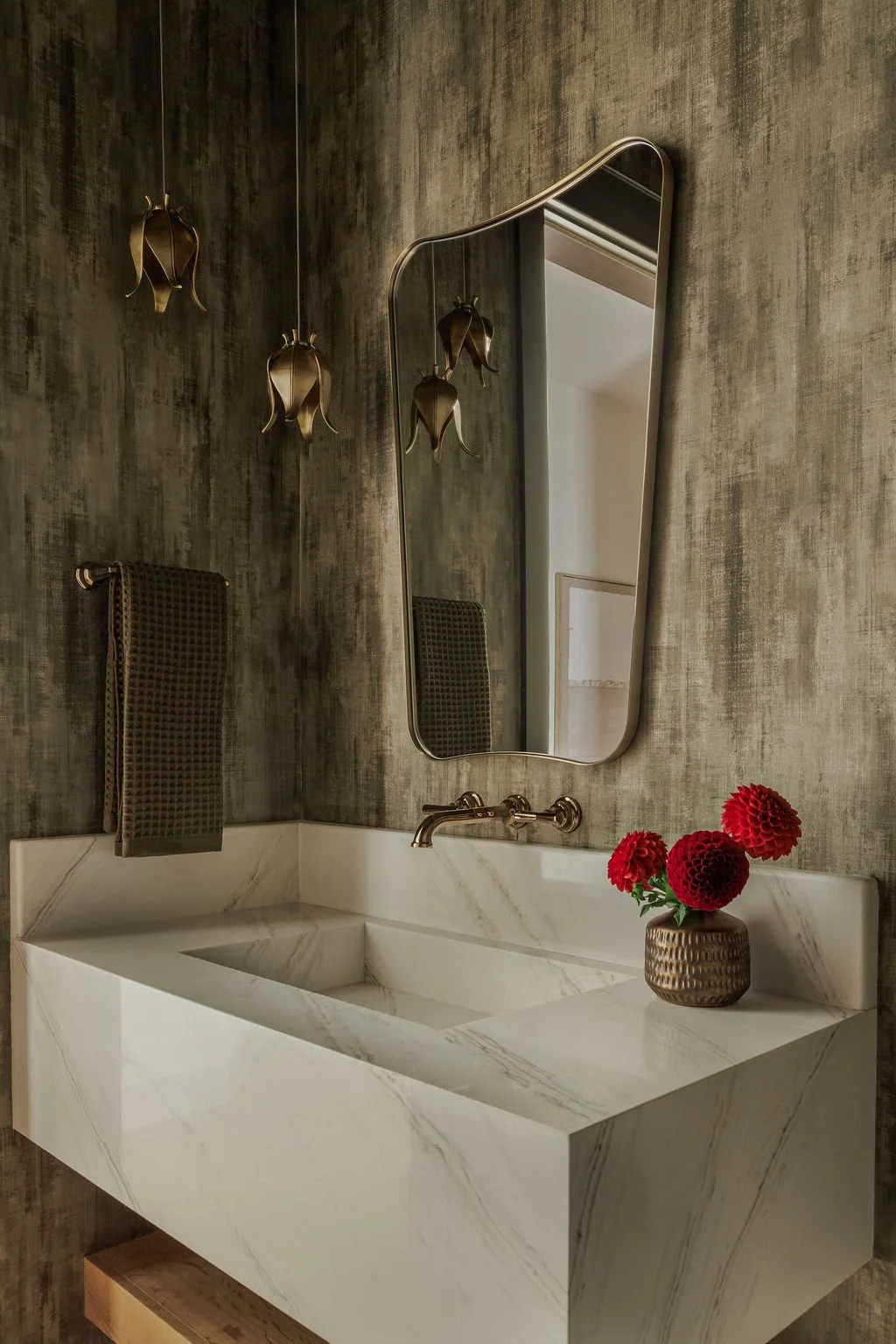 Modern bathroom vanity with a white marble sink, a gold faucet, a round mirror, a vase with red flowers, a towel, and decorative hanging sconces on textured wall.