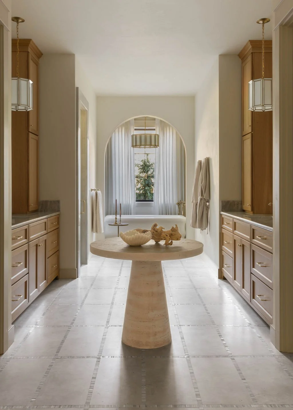 A bright kitchen with beige cabinets, a central round stone table with bowls and decorative objects, and an arched opening leading to a bright room with a window and sheer curtains.